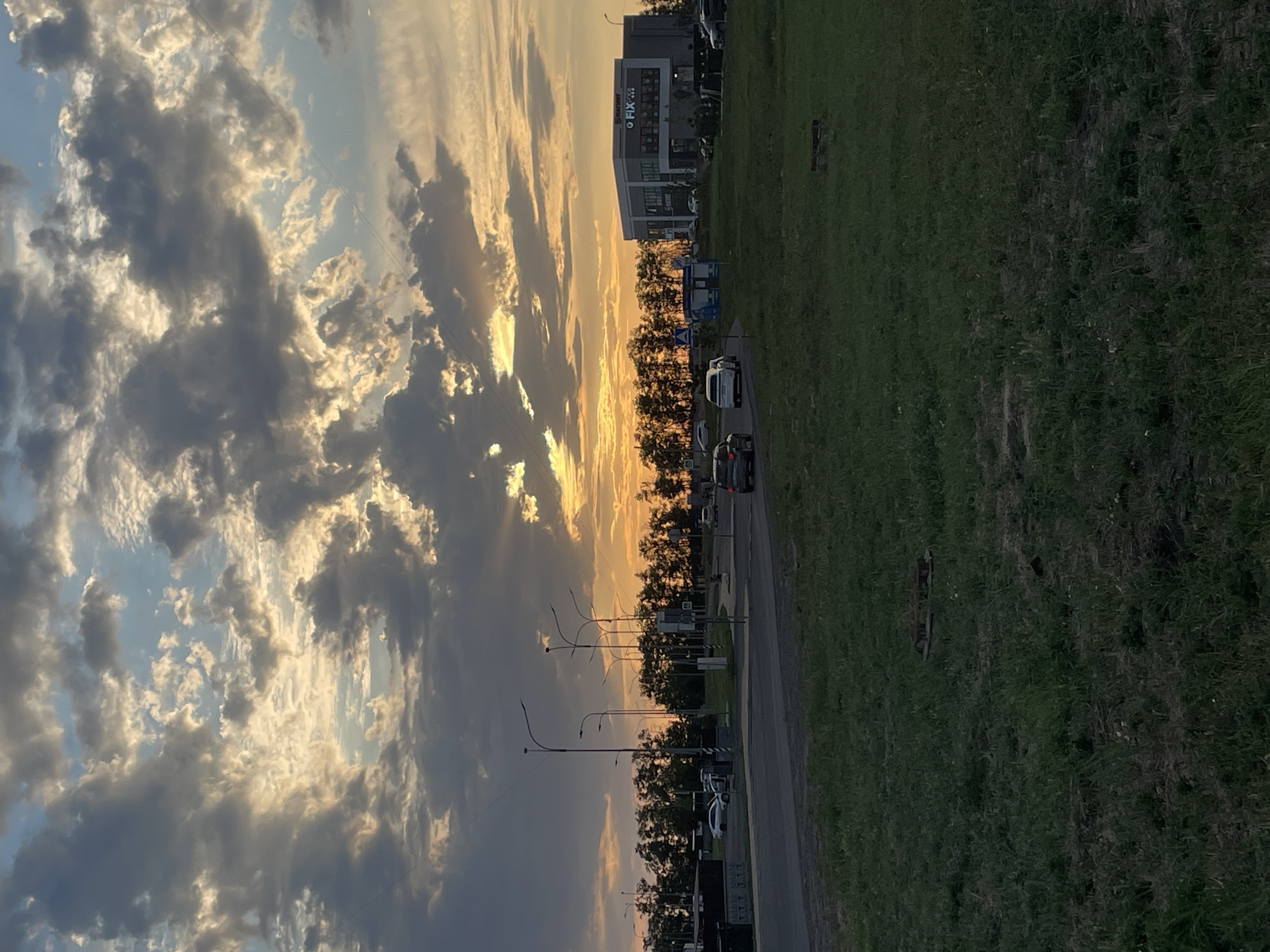 The image captures a suburban scene in Rostov-on-Don, Russia, during sunset. The sky is filled with dramatic clouds illuminated by the setting sun, casting a warm glow on the landscape. In the foreground, a patch of grass is visible, with a manhole cover and a wooden bench scattered across it. A paved road curves through the middle ground, with several cars driving on it. A line of trees forms a dense backdrop behind the road. On the right side of the image, a modern, two-story building with a sign that reads "FIX PRICE" stands out. Streetlights dot the area, and power lines crisscross the sky. The overall impression is one of a typical evening on the outskirts of a city.