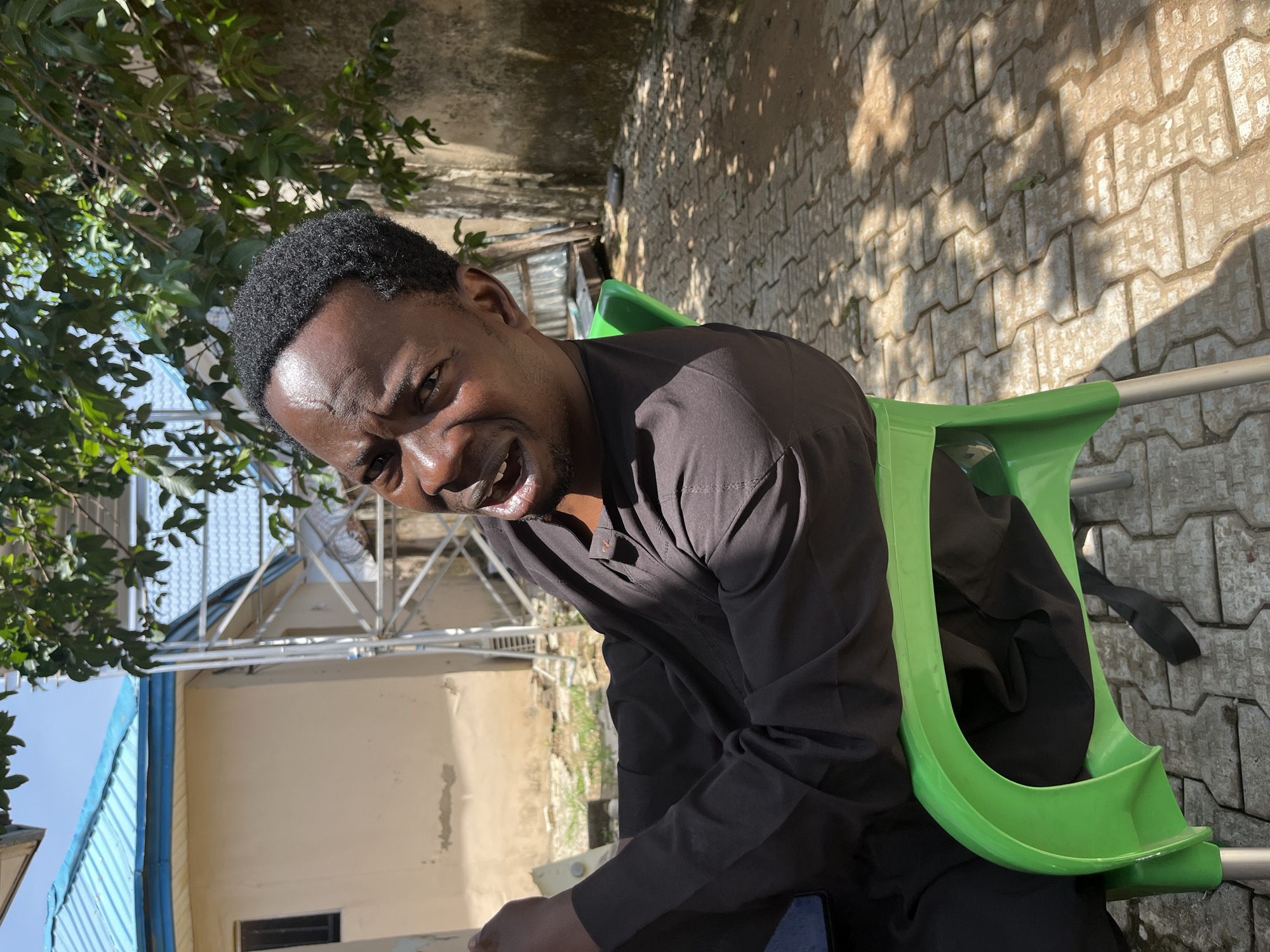 A man with dark skin and short, dark hair is sitting on a bright green plastic chair outdoors. He is wearing a dark, possibly black, long-sleeved outfit with a buttoned collar. His head is tilted, and he is looking directly at the camera with a wide, open-mouthed expression, suggesting he is speaking or laughing. Sunlight is casting shadows on the textured paving stones behind him, indicating it is daytime. Lush green foliage from a tree is visible on the left side of the frame, partially obscuring a building in the background. The building appears to be light-colored with a blue roof. The overall scene is casual and appears to be in a residential or public outdoor area.