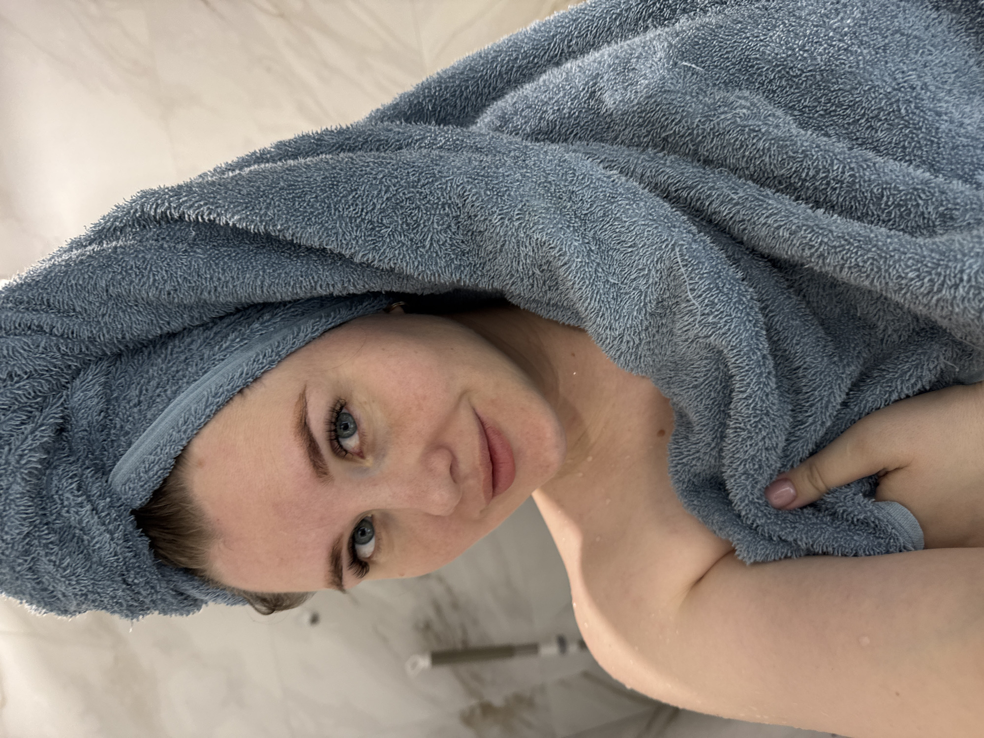 A woman is shown from the chest up, wrapped in a blue towel on her head and body. There are visible water droplets on her chest and shoulder. The background appears to be a bathroom with light-colored marble or tile. The location is unknown but is asked to be in Gornyi (Dvurechenskoe S/P), Russia.