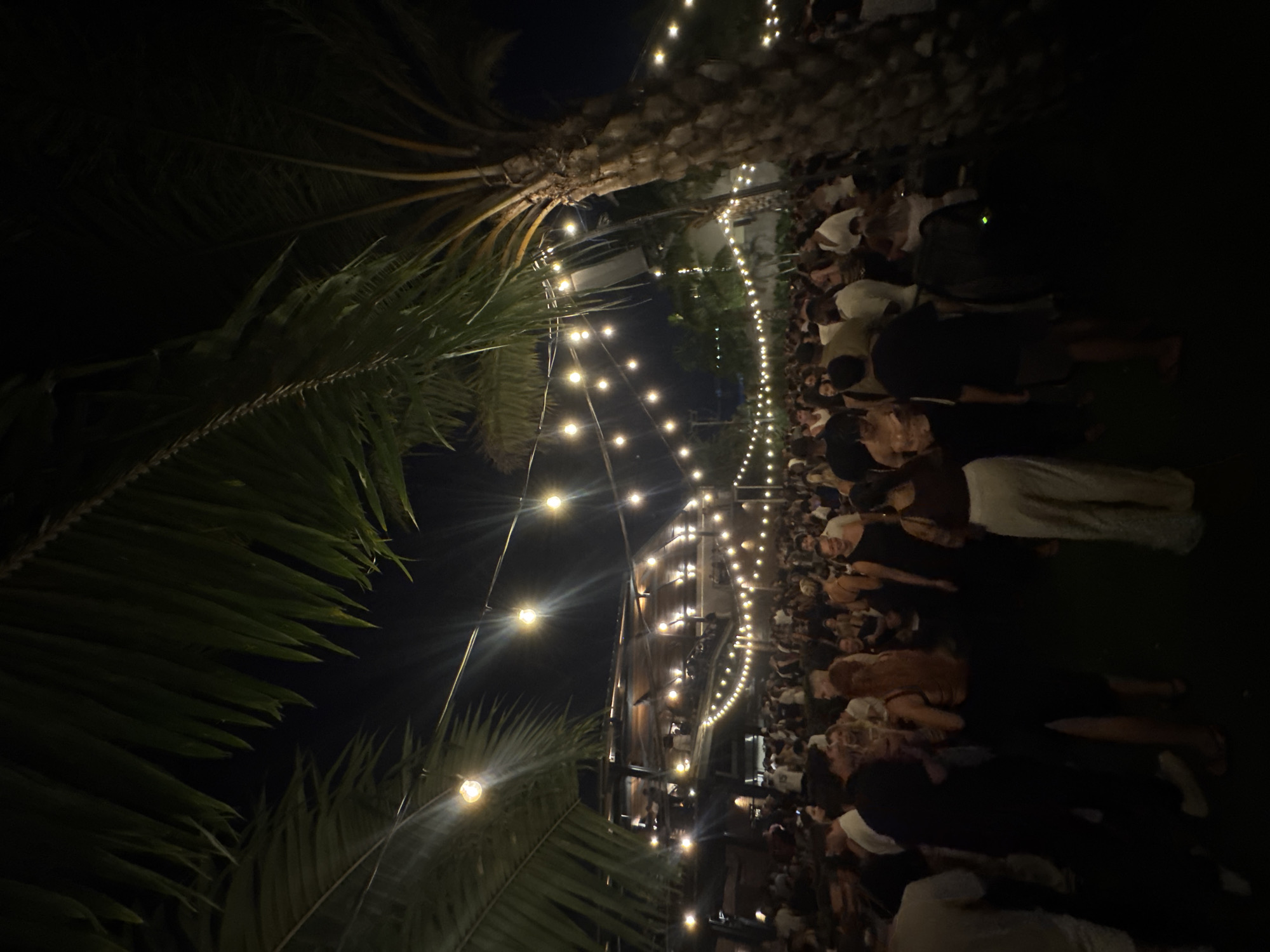 A nighttime outdoor gathering under string lights is taking place in what appears to be North Kuta, Indonesia, indicated by the presence of palm trees. A large group of people are standing closely together. The scene is mostly dark with light provided by the string lights and some ambient lighting.
