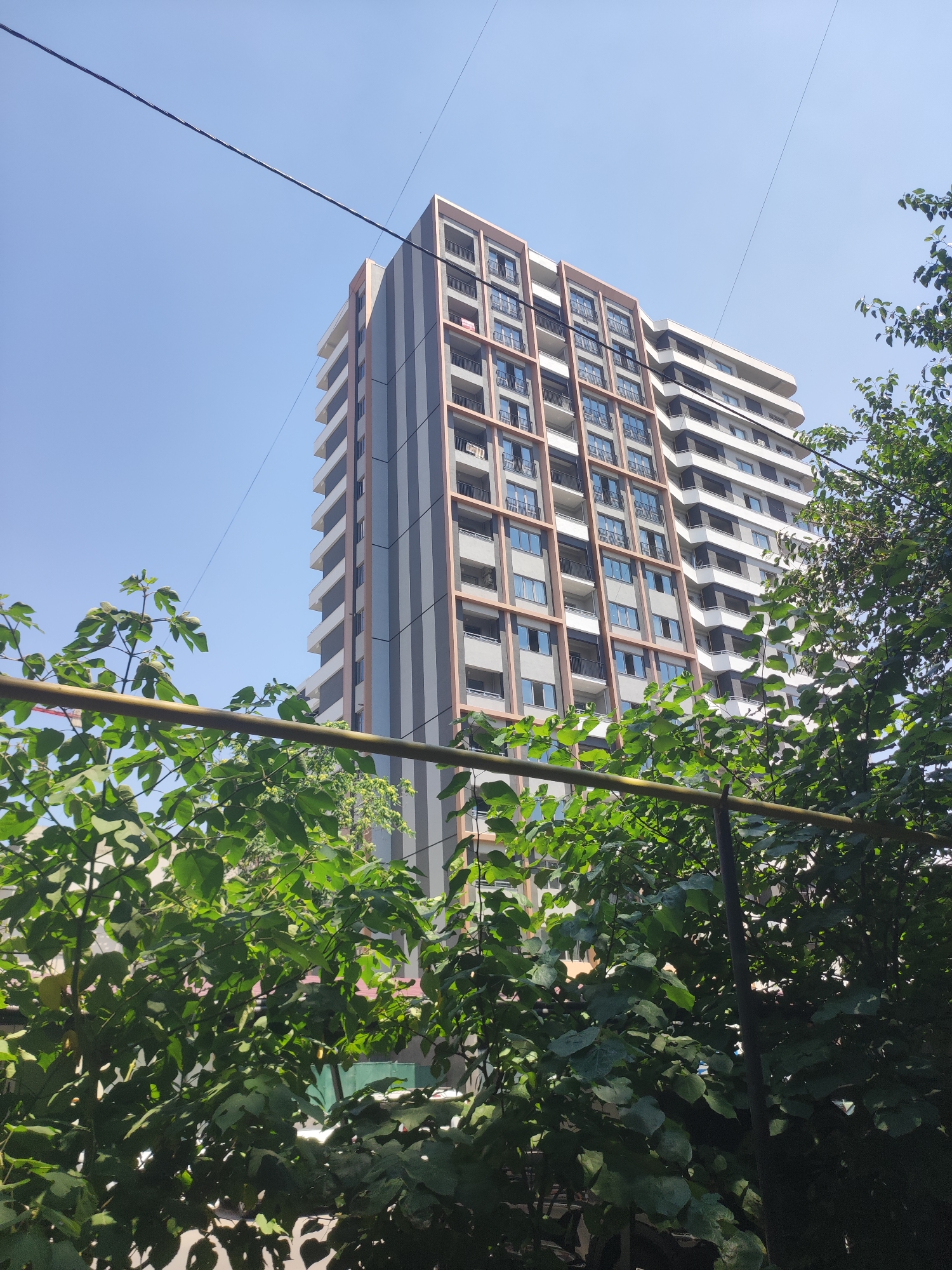 The image shows a tall, modern apartment building in Tashkent, Uzbekistan. The building has a distinct architectural design with alternating vertical bands of gray and a reddish-brown color, interspersed with rows of balconies. The sky above is clear and blue. In the foreground, lush green foliage obscures the lower portion of the image, with a horizontal metal bar running across. Overhead power lines are visible against the sky, stretching across the frame and partially in front of the building. The scene suggests a sunny day in an urban environment.