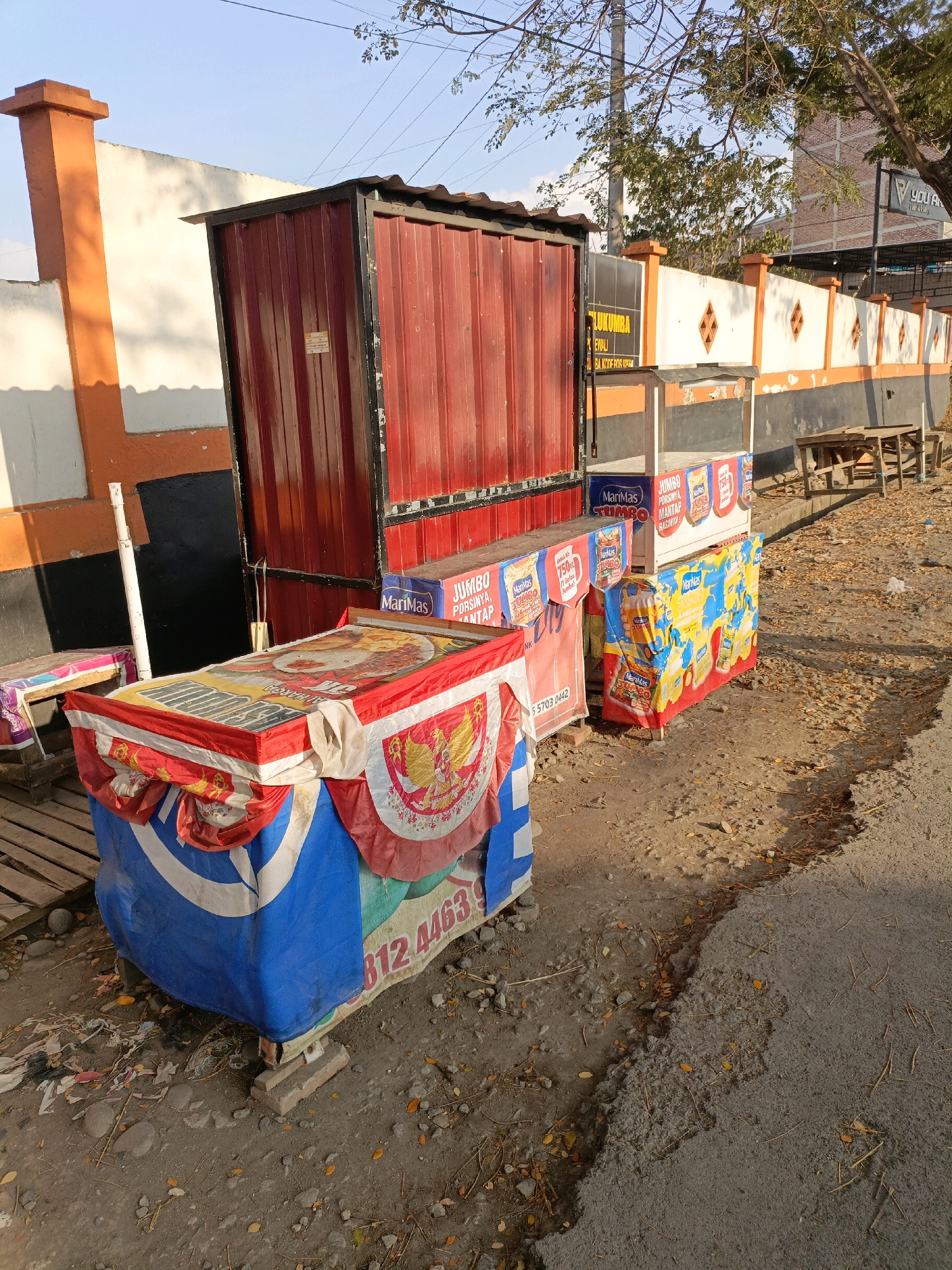 The image captures an outdoor scene in Bulukumba, Indonesia, characterized by a street-side food stall. The stall features a prominent red shipping container-style structure serving as a backdrop, with multiple smaller display units in front. These display units are covered with colorful banners advertising various food products, including what appear to be snacks and drinks. Notably, one banner displays the Indonesian coat of arms, Garuda Pancasila. Several stacks of processed food packages, some labeled "Jumbo" and "MariMas," are visible. The ground is a mix of dirt, gravel, and broken asphalt, with scattered leaves and debris, indicating a potentially unpaved or neglected area. A simple wooden bench is visible in the background, alongside a wall with orange and white paint. The lighting suggests it is late afternoon, with long shadows cast by the structures. No people are present in the scene.