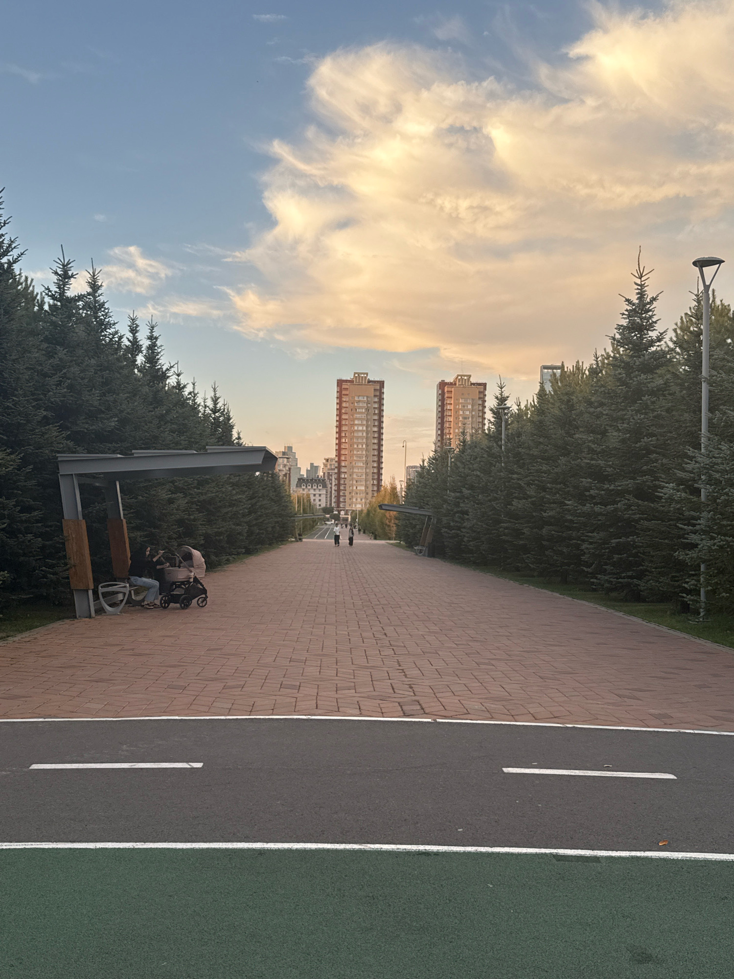 The image captures a scenic view of a park pathway in Astana, Kazakhstan, during what appears to be the late afternoon or early evening, given the warm lighting and the soft hues of the sky. The pathway itself is constructed of red brick pavers, forming a wide, straight avenue that stretches towards the horizon. To the left and right of the brick pathway, dense rows of evergreen trees, likely spruce or fir, create a natural border, with their branches arching slightly inwards.
In the foreground, a distinct asphalt strip with white line markings indicates a bike lane or a running track, separated from the pedestrian path by a white boundary line. Further into the scene, a modern architectural element, possibly a bus shelter or a pavilion, is visible on the left side. Underneath this structure, a person is seated on a bench next to a baby stroller, suggesting a moment of rest or observation.
Dominating the background are several tall, modern residential buildings with distinctive architectural designs, their facades catching the golden light of the setting sun. The sky above is a canvas of soft blue with large, fluffy clouds tinged with shades of yellow and orange, indicative of sunset. A lamppost stands tall on the right side of the pathway, adding to the urban park setting. In the distance, a few individuals are walking along the path, adding a sense of scale and life to the scene. The overall impression is one of a well-maintained urban green space designed for recreation and relaxation.