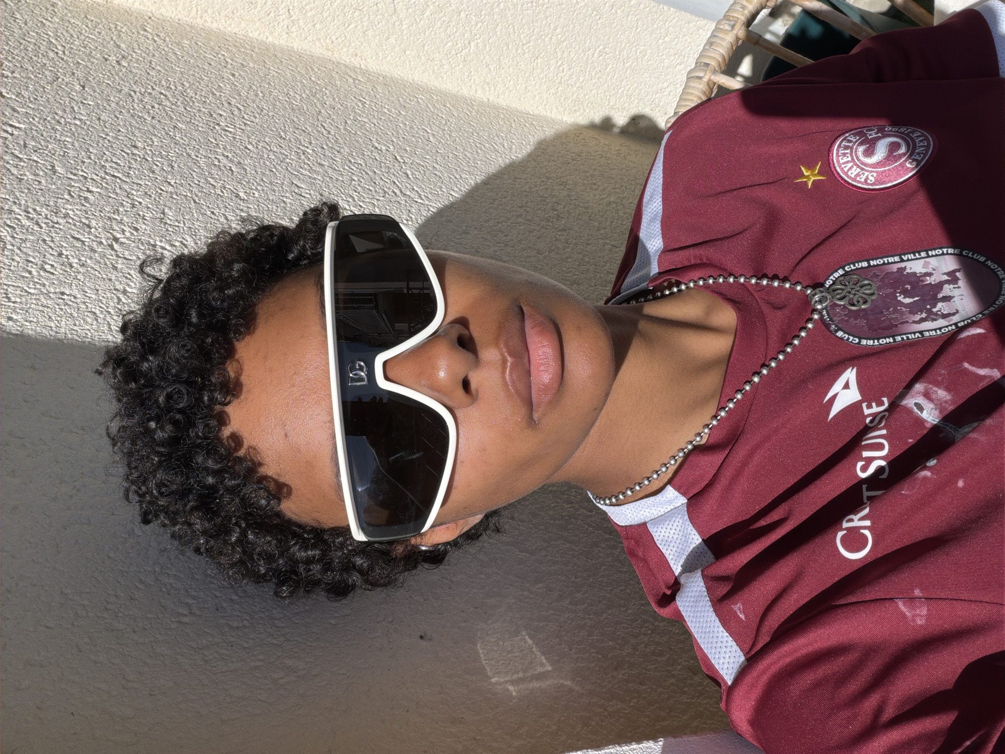 A person with dark curly hair and brown skin wears large, white-framed sunglasses and a maroon soccer jersey, lying against a textured white wall. The jersey features a white logo that reads "CRT SUISE", the Servette FC team badge, and a beaded necklace. The scene is bathed in sunlight.