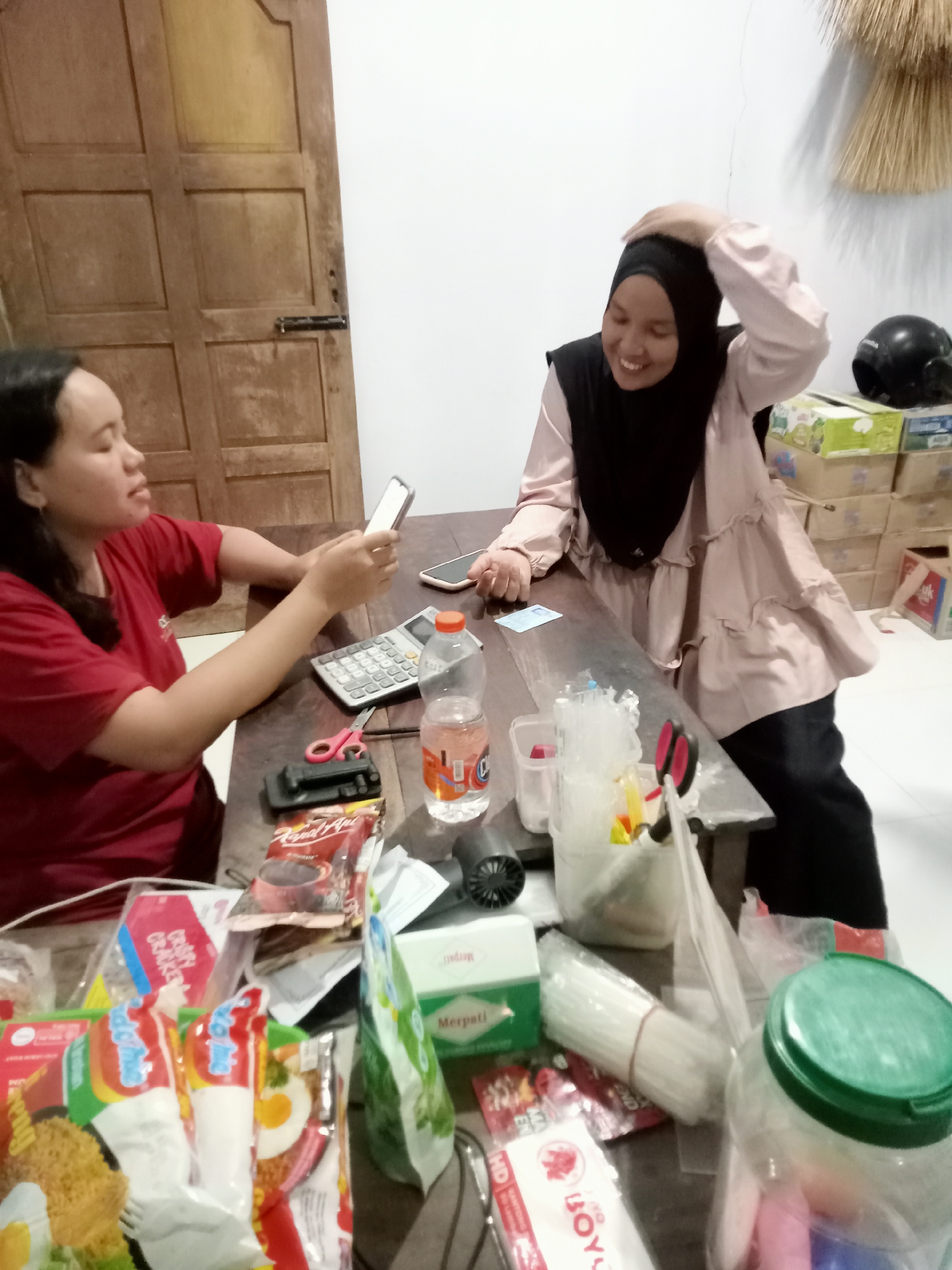 The image depicts an indoor scene, possibly a home or small shop in Simo, Indonesia, with two women interacting around a wooden table laden with various items. On the left, a woman wearing a red t-shirt is seated and holding a smartphone, seemingly looking at it. The woman on the right is wearing a hijab and a light pink ruffled blouse, and she has her hand on her head, appearing to be laughing or smiling.
The table is cluttered with an assortment of objects, suggesting a casual or perhaps busy environment. Among the items visible are a calculator, scissors, packets of instant noodles and snacks, a bottle of water, a plastic container filled with what appear to be straws or similar items, and a large glass jar with a green lid. In the background, a wooden door with a metal latch is visible, and stacks of cardboard boxes are piled against a white wall. A black motorcycle helmet is also present in the background, near the boxes. The lighting in the room appears to be artificial and somewhat dim.