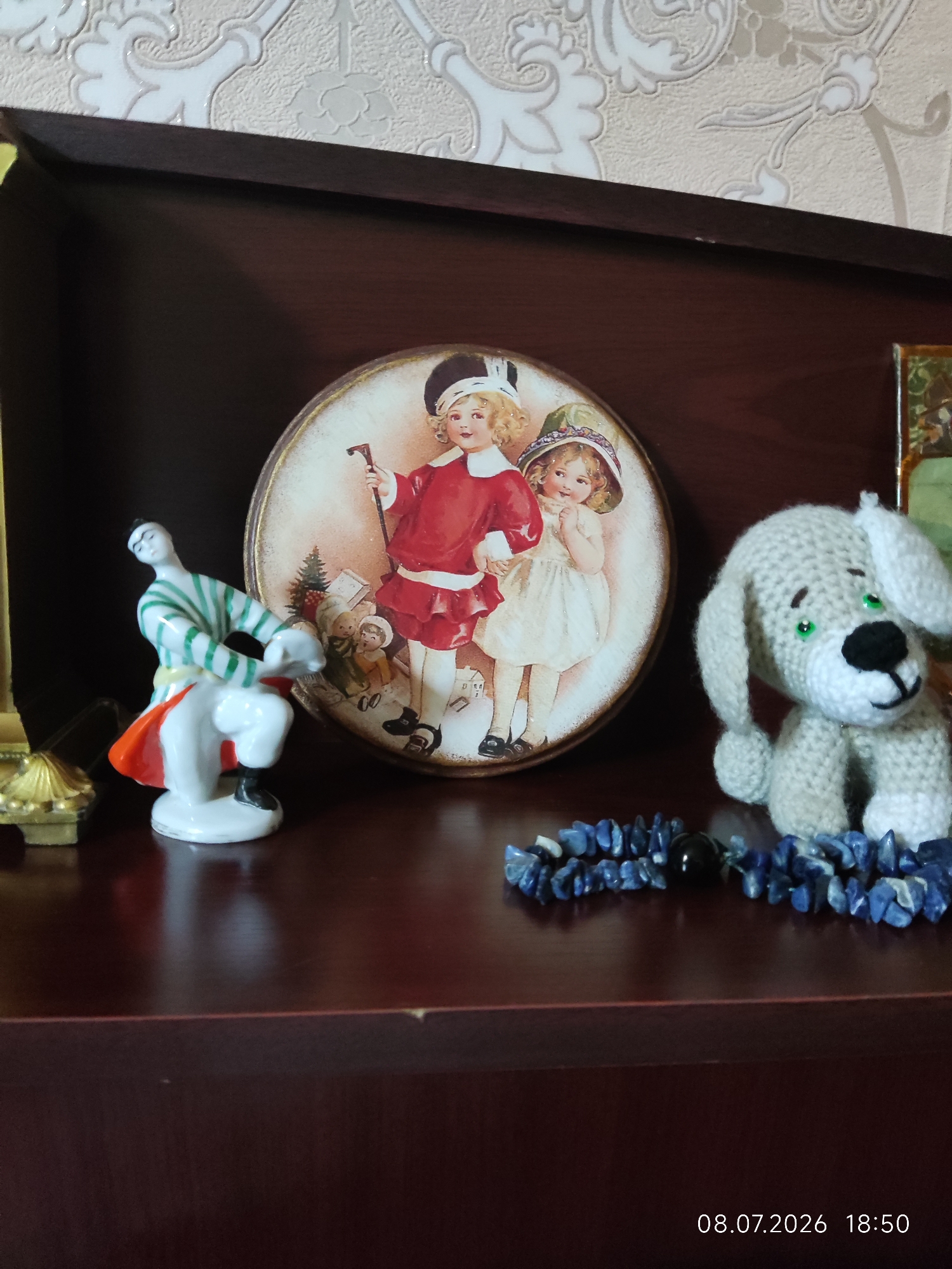 A shelf in Karshi City, Uzbekistan, displays several decorative items. On the left, a porcelain figurine of a person in a striped green and white shirt, white pants, and an orange skirt is posed in a dynamic stance. To its right, a round decorative plate features an illustration of two young children, one in a red outfit holding a cane and the other in a white dress. A crocheted gray dog with green eyes and a black nose sits to the right of the plate. In front of the dog is a blue beaded bracelet with a single black bead. The shelf is made of dark wood, and a light-colored patterned wallpaper is visible in the background. A timestamp in the lower right corner reads "08.07.2026 18:50".