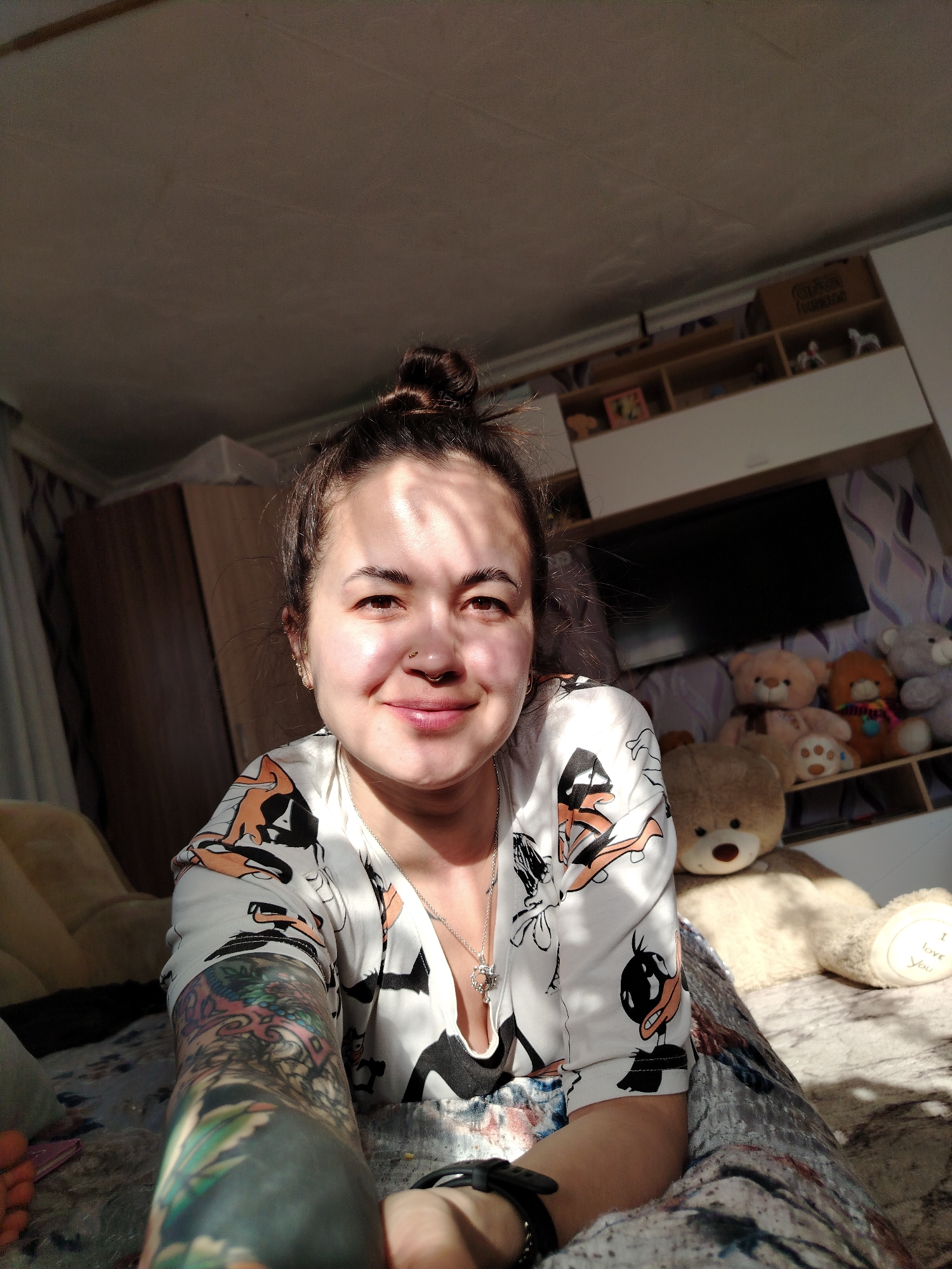 A woman with brown hair in a high bun and a nose piercing is shown in a medium close-up selfie. She is smiling and wearing a white shirt with cartoon character prints, a silver necklace, and a watch on her left wrist. Her left arm is covered with a detailed tattoo.
Behind her is a bed covered with a patterned blanket. On the bed is a large teddy bear. Behind the teddy bear are several other teddy bears and a flat-screen television mounted on a wall. A wooden cabinet with shelves is above the television. A partial view of a wooden wardrobe can be seen to the left. The room is lit with natural light, which casts shadows across the woman's face. The location appears to be an interior residential space, most likely in the city of Гожан, Russia.