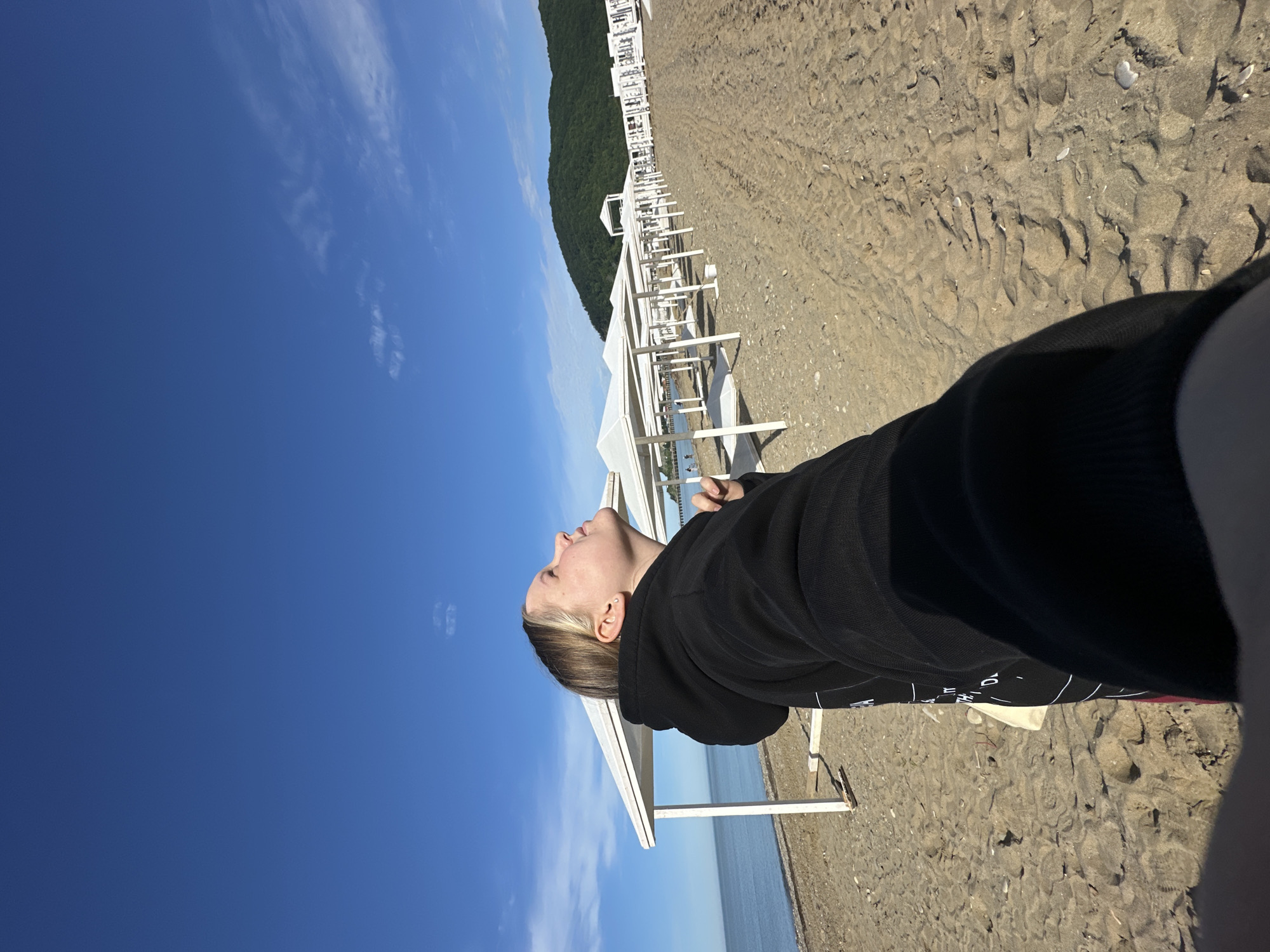 An outdoor, eye-level shot captures a sunny day on a beach. In the foreground and on the left, a person, possibly female, is seen in profile, looking upwards with their head slightly tilted back and eyes appearing closed or focused on the sky. They have light-colored hair, possibly blonde or light brown, and are wearing a black hooded sweatshirt with some faint white text and lines visible on the lower front/side. A small stud earring is visible in their ear.
The background reveals a wide, sandy beach stretching into the distance on the right, with distinct tire tracks and small shells scattered on the sand. A long row of white, open-sided beach shelters or cabanas extends across the mid-ground, leading towards the horizon. Beyond these structures, a calm blue sea is visible. In the far background, a large, dark green, forested mountain or hill rises prominently, indicating a coastal mountain region. The sky above is a clear, vibrant blue with a few wispy white clouds. The overall scene suggests a peaceful, relaxing moment on a beach, likely in Lermontovo, Russia, during daytime hours on a clear and sunny day.