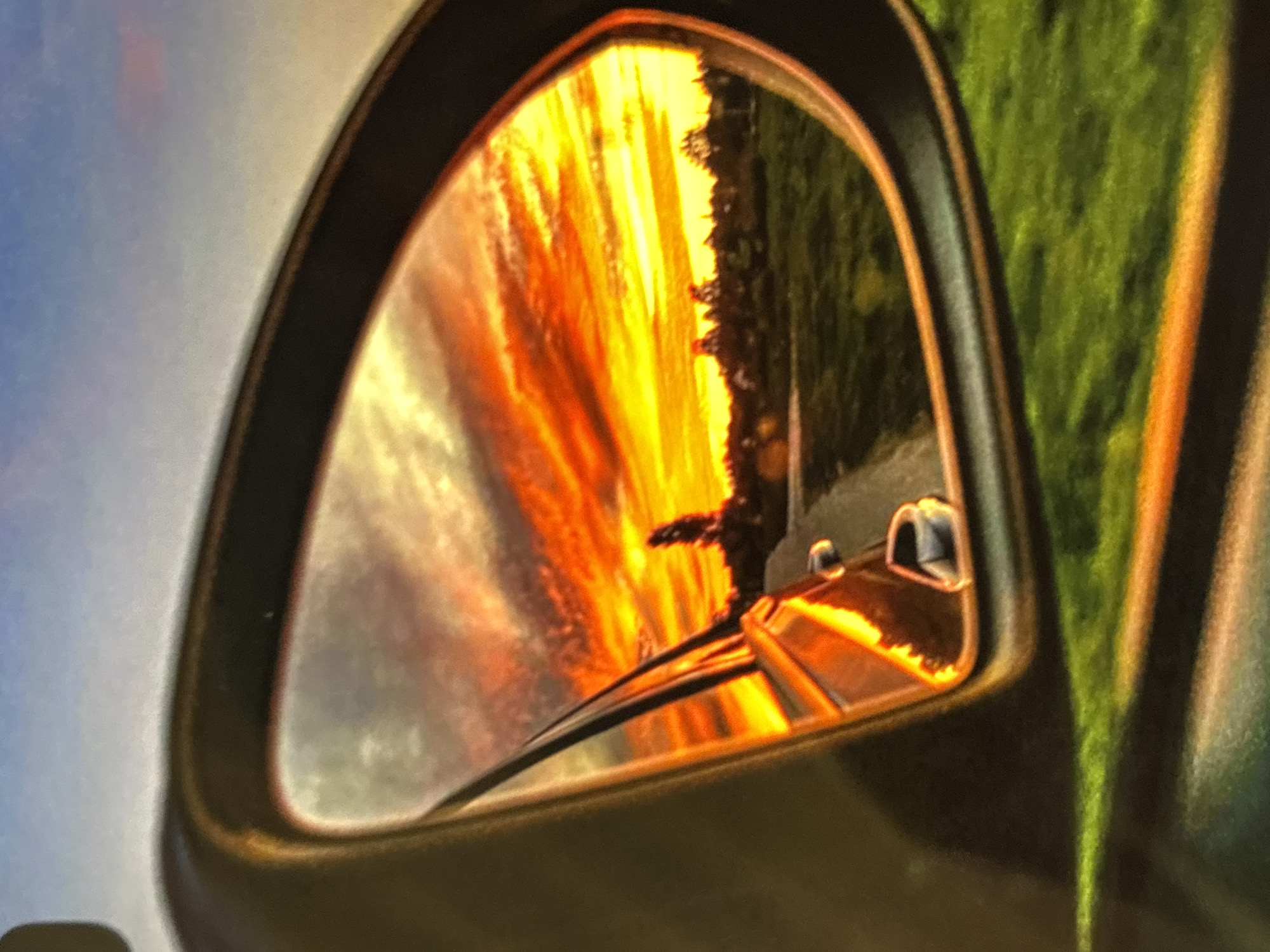 The image shows a car's side mirror reflecting a vibrant sunset. The sunset colors include oranges, reds, and yellows, blended with clouds. The reflection also shows a road and some trees lining the side. Part of the car, specifically the wiper blades and a portion of the dashboard, is visible in the mirror's reflection. The rest of the car and the surrounding environment is out of focus. I have no information about Belopoptsi, Bulgaria, or the image's location.