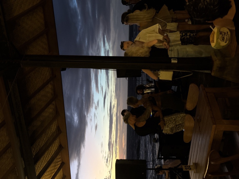 This image depicts an outdoor, beachfront social gathering at "Sunset Point" in Canggu, Indonesia, during twilight or dusk. The scene is observed from under a thatched roof structure, with a prominent dark support pillar dividing the view.
In the midground, several individuals are present. To the left of the pillar, a couple is dancing intimately near a DJ setup, which includes two large black speakers and a dark table. The man, with light hair and visible arm tattoos, wears a black sleeveless top and patterned trousers. The woman faces him, her back to the viewer, wearing a dark top. Behind them, the Indian Ocean is visible with waves, under a sky displaying a gradient from pale orange-yellow at the horizon to dark blue-purple with extensive cloud cover.
To the right of the pillar, a group of people are standing and socializing. A man with a beard and sunglasses wears a light-colored short-sleeved shirt and patterned shorts. A woman with long blonde hair is partially obscured by the pillar, and another woman with long blonde hair in a light-colored dress stands further right.
In the immediate foreground, a wooden table with chairs is partially visible on the left, featuring a small yellow sign with the number "22" affixed to its leg. On the right, at the base of the pillar, a coconut drink with a straw and a "Sunset Point" logo is visible. A person's head and shoulder are visible in the lower right, looking towards the scene. Artificial lighting illuminates the area under the roof structure.