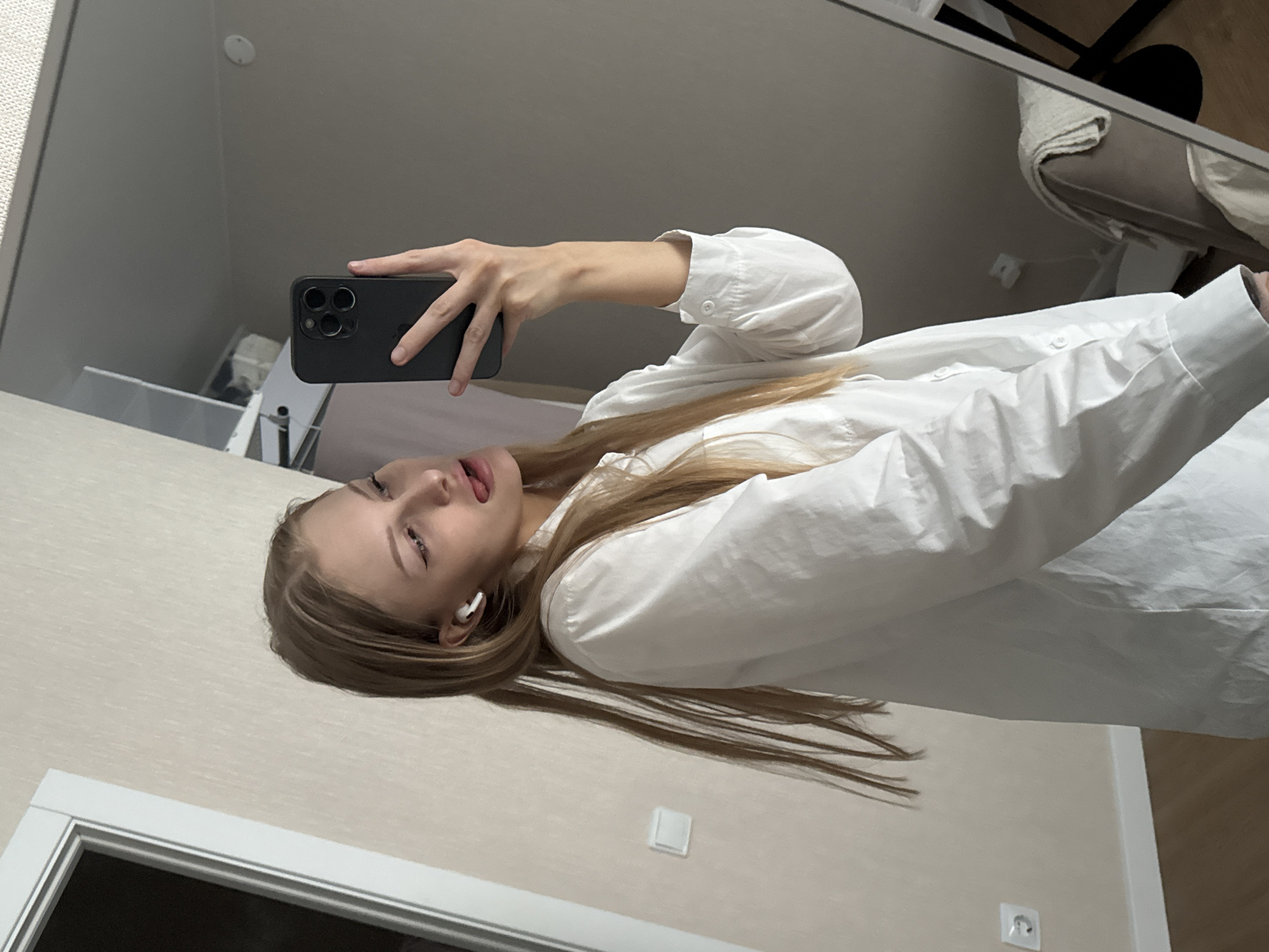 A young woman with blonde hair is taking a selfie in a room, possibly a bedroom or dressing area. She is wearing a white button-down shirt and white wireless earbuds. Her tongue is sticking out playfully. She is holding a black smartphone in her right hand, which is angled to capture her reflection in a mirror. The background includes a pale, textured wall with a light switch, a portion of a doorframe, and a hint of a mirror reflecting furniture and a blanket. The overall impression is casual and informal. This image was captured in Perm, Russia.