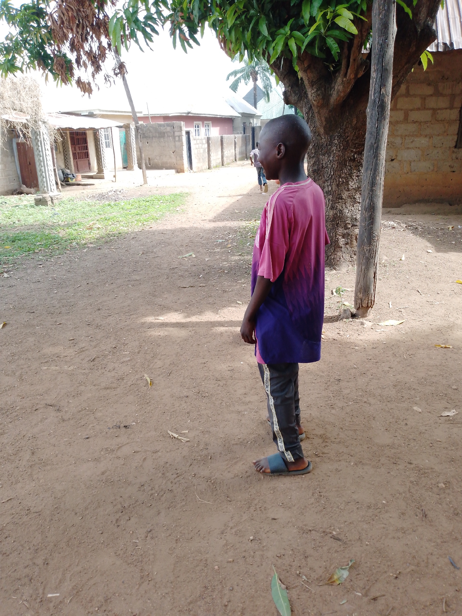 An outdoor, eye-level shot captures a young person standing on an unpaved dirt path in what appears to be a village or rural settlement. The person, viewed from behind and slightly to their left, has dark skin and a closely shaven head. They wear a short-sleeved t-shirt that transitions from pink at the top to purple at the bottom, and dark trousers with a white vertical stripe featuring repeating text, partially visible as "ATMOSPHERE". Dark slides or sandals are on their feet. The person's head is turned to the left, and a hint of a smile is visible on the left side of their face.
To the right, a large tree with a thick, textured trunk and green leaves dominates the frame. A thin, light-colored wooden pole stands upright near the tree. The ground is dry, light brown earth with scattered leaves and small patches of green grass. In the background, several simple buildings are visible, constructed from concrete blocks, some with light-colored walls and corrugated iron roofs. One building has a reddish-brown door, another has white-framed windows. Faintly in the distance, another person can be seen walking away. The scene is brightly lit by natural sunlight, suggesting daytime, likely late morning or early afternoon, with shadows cast on the ground. The overall setting implies a warm climate and a simple, residential environment.