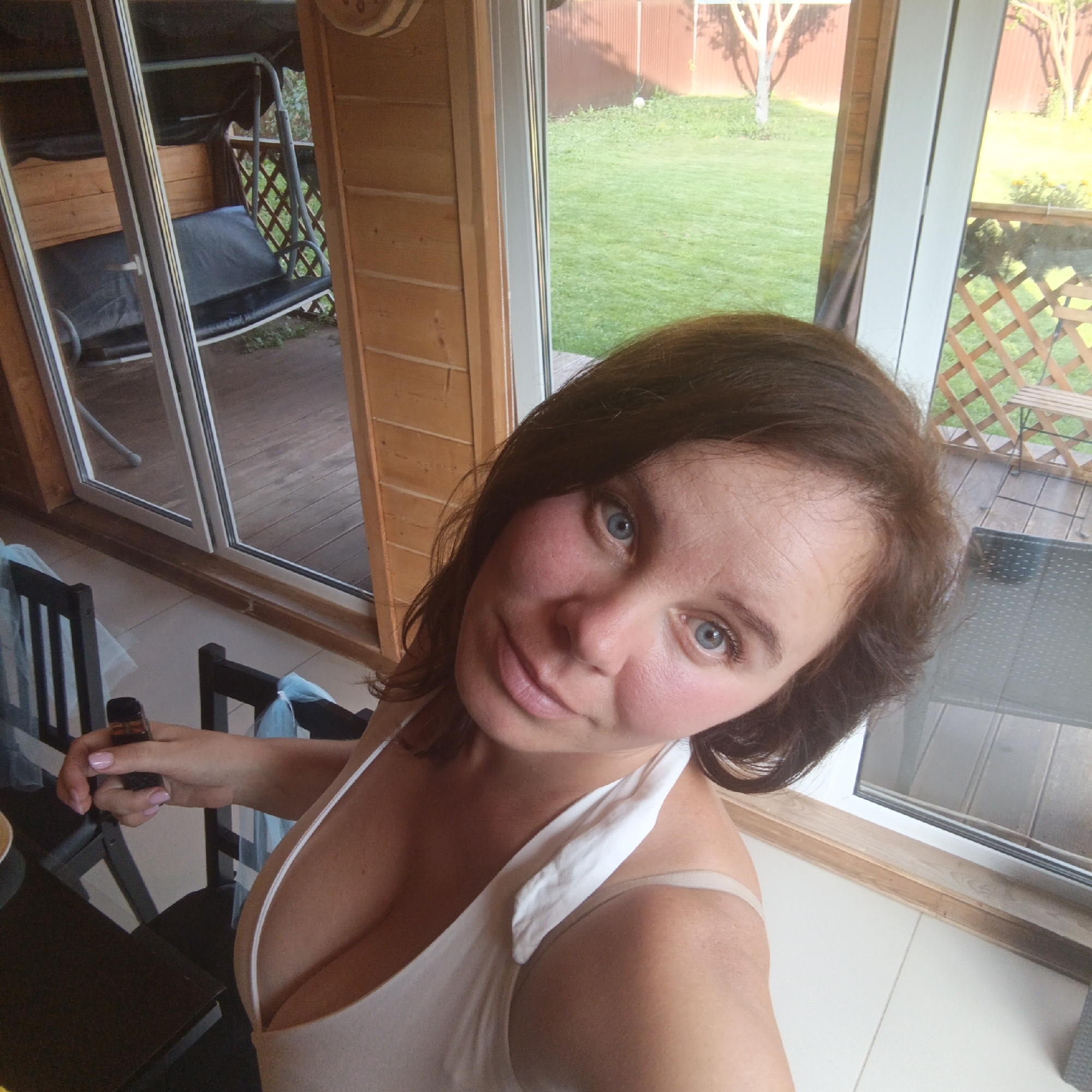 An adult Caucasian female with brown hair and blue eyes is positioned in the foreground of a domestic setting, possibly a veranda or sunroom, in Chaikovsky, Russia. She is wearing a white tank top. Her right hand is holding a dark-colored object, possibly a vape pen. She is looking at the camera, with her head tilted.
The background reveals a large glass door and window that looks out onto a grassy yard with a wooden fence and trees. Inside, wooden paneling lines the walls, and a portion of a black chair is visible to the left of the woman. A suspended swing is visible through the glass door on the left. The flooring appears to be light-colored tile. The overall impression is a casual, indoor-outdoor living space.