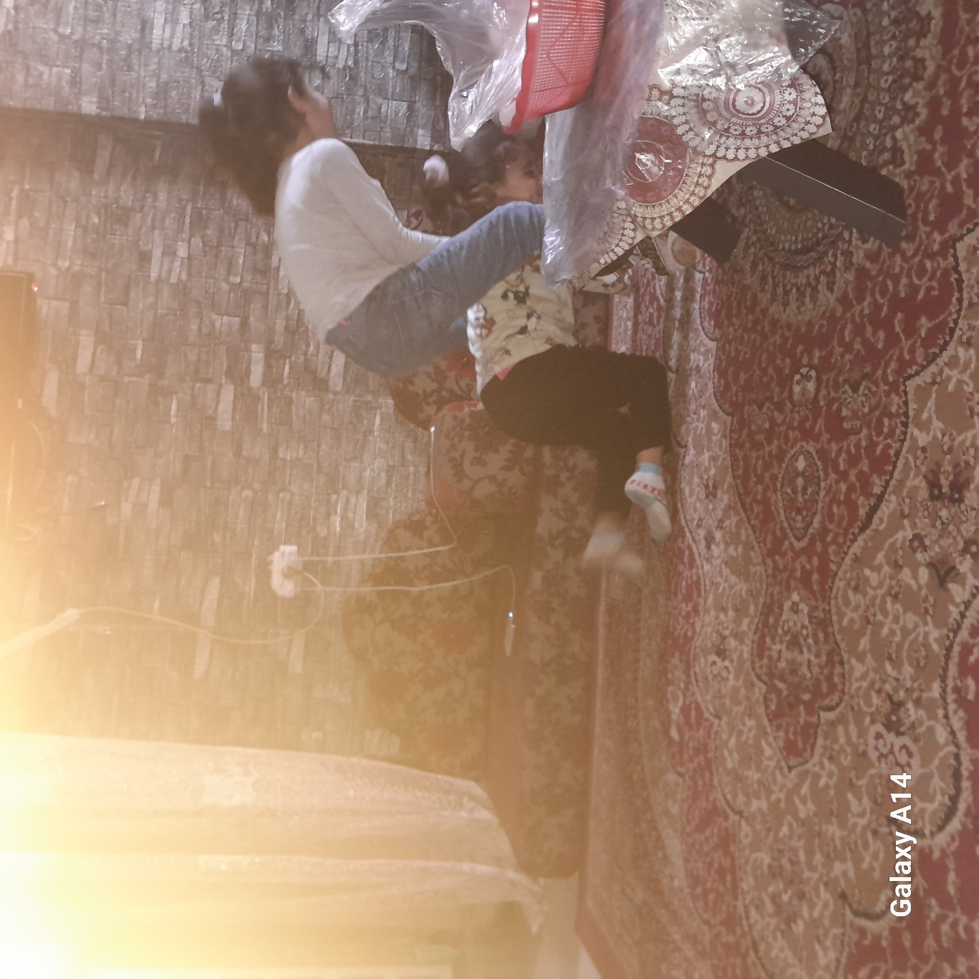 Two children are present in an indoor setting. One child is wearing a white long-sleeve shirt and jeans, and the other child is wearing a patterned shirt and black pants. The children are positioned near a table with a patterned tablecloth and a red basket covered in plastic. The environment features a brick-patterned wall and a patterned carpet. The city and specific location are unknown.