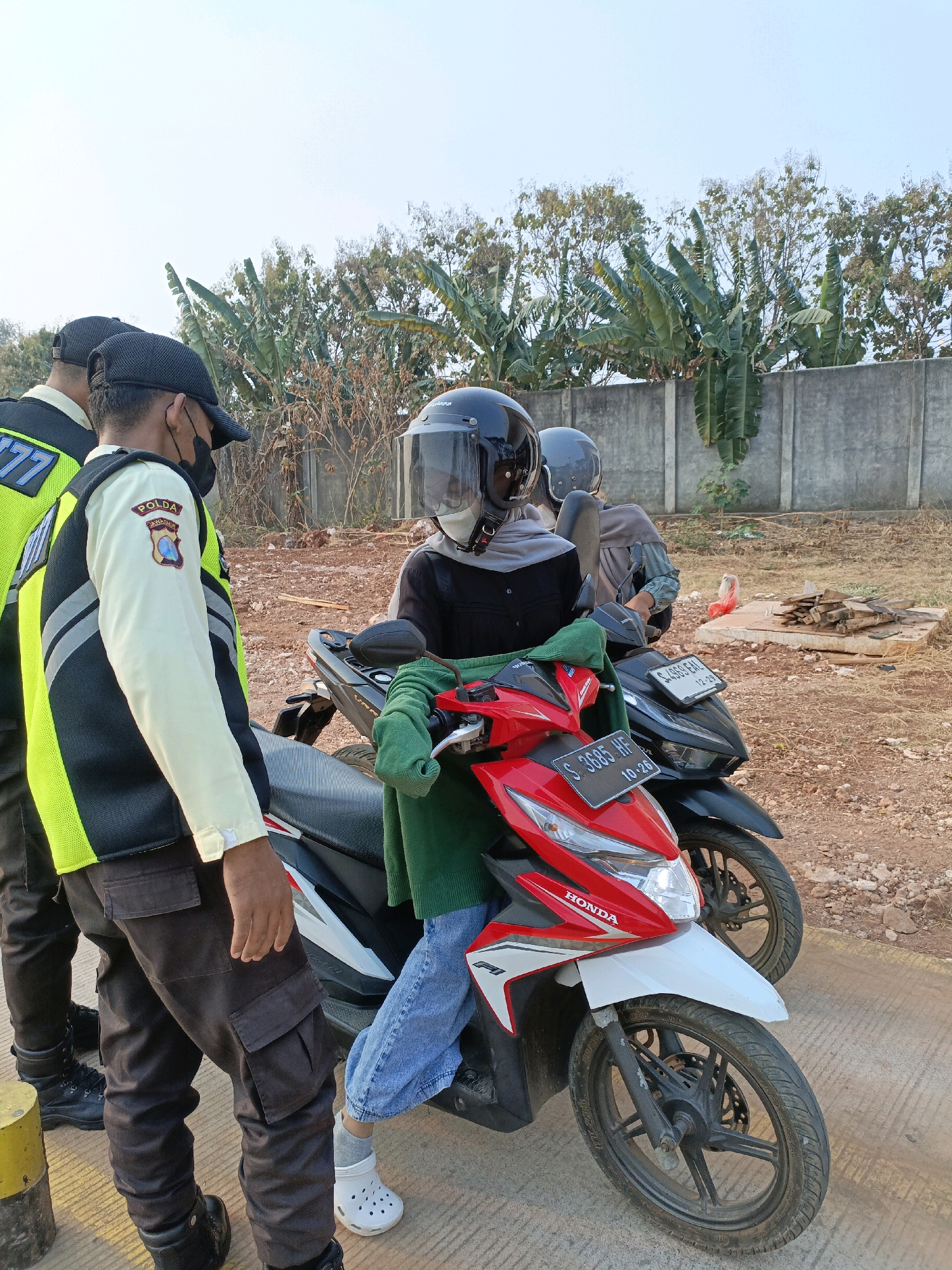 In Jenu, Indonesia, two uniformed personnel, likely police officers based on their reflective vests and caps, are interacting with two individuals on motorcycles. The officers are positioned on the left side of the frame, one facing away from the camera and the other partially turned. They wear dark pants and vests with reflective yellow and black detailing, and one has a badge on his arm with "POLDA JAWA TIMUR" visible. The two individuals on motorcycles are wearing helmets and face coverings. The person on the red and white scooter in the foreground is wearing a green garment and blue jeans, while the person on the black scooter behind them wears a gray garment. Both scooters are parked side-by-side. The environment appears to be an outdoor area with a dirt and gravel ground, a concrete wall in the background, and trees, including banana trees, visible behind the wall. The sky is clear and bright.
