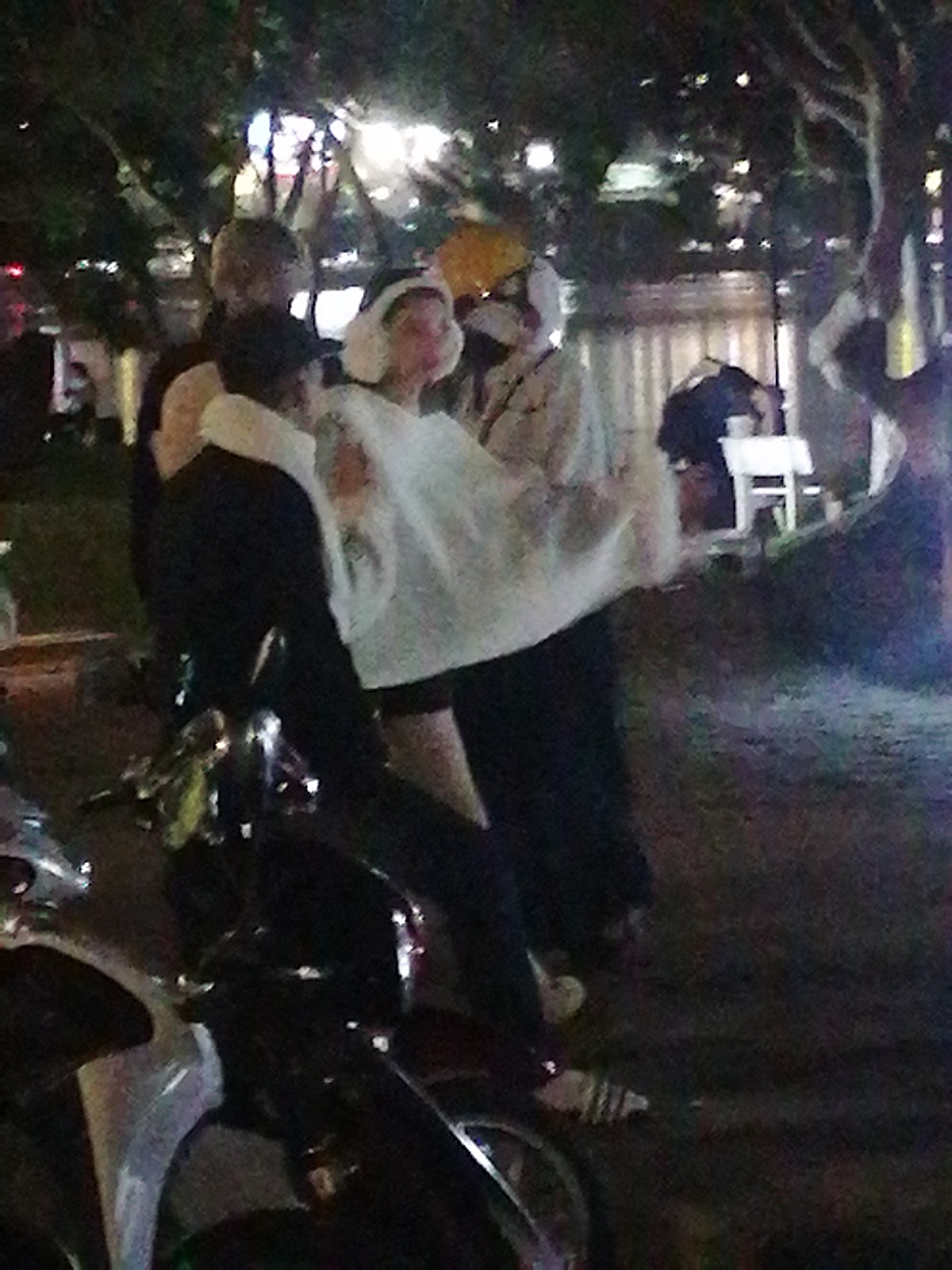 In Da Lat, Vietnam, at night, a group of people are gathered outdoors, possibly in a park or on a street with trees. In the foreground, a motorcycle is partially visible. A person in the center is wearing a white, loose-fitting garment that appears to be draped over them, possibly a blanket or a large piece of fabric. They are also wearing a white head covering. To the left, another person is wearing a black jacket. In the background, the scene is illuminated by artificial lights, creating a somewhat blurry and dimly lit atmosphere. There are hints of other individuals and structures in the background, suggesting a public area.