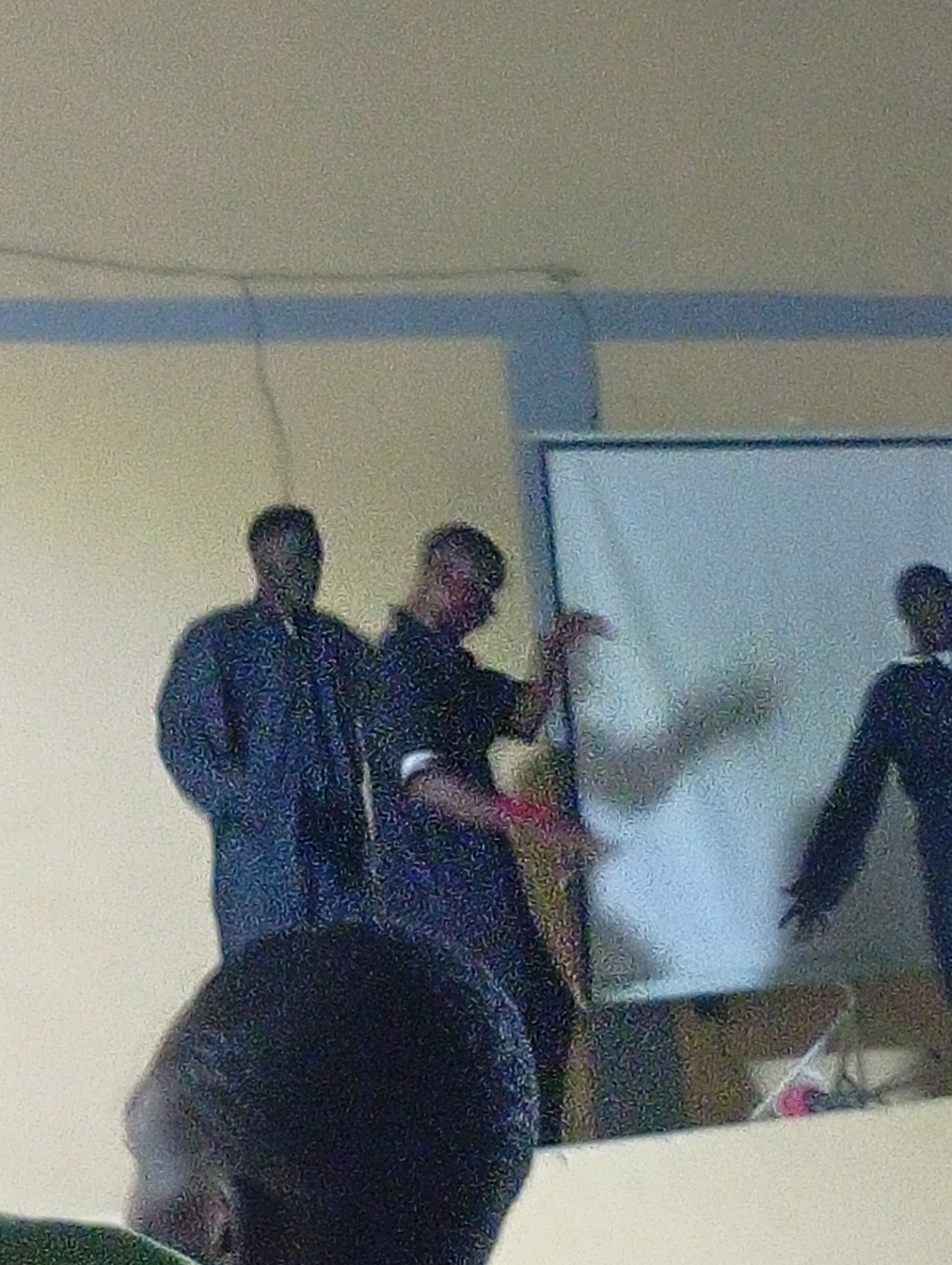 The image shows three people standing in front of a white projection screen, which is framed in blue. The setting is a room with beige walls and a white ceiling, presumably indoors. The location in Maiduguri, Nigeria is unable to be determined based on the image. The people are wearing dark clothing, and one individual is gesturing with their hands toward the screen. Another person stands slightly behind and to the left of the gesturing individual. A third person stands on the right side of the projection screen. There is a partial view of a person's head in the foreground. There's an indistinguishable object on the stand below the projection screen.