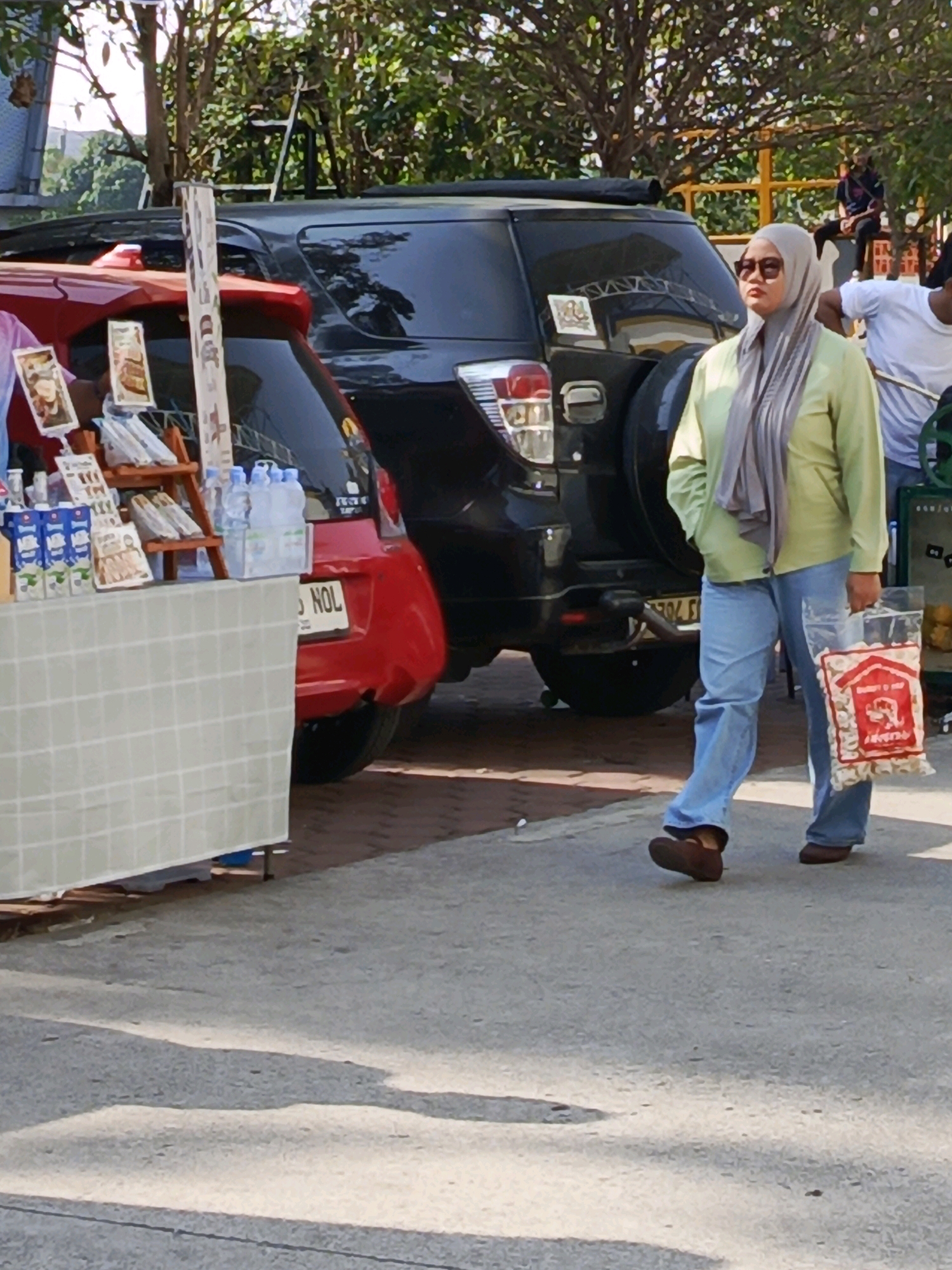 A woman wearing a grey hijab, light green long-sleeved shirt, and blue jeans is walking on a paved surface. She is carrying a clear plastic bag filled with small, red and white patterned items. She is wearing dark sunglasses and brown shoes. To her left is a red car with a license plate that reads "5 NOL". Behind the red car is a dark SUV with a spare tire mounted on the back. A table covered with a checkered cloth is in the foreground. The scene appears to be outdoors, possibly a street market, given the presence of the table with items and vehicles. The lighting suggests it is daytime. The environment includes trees and sunlight creating shadows on the ground.