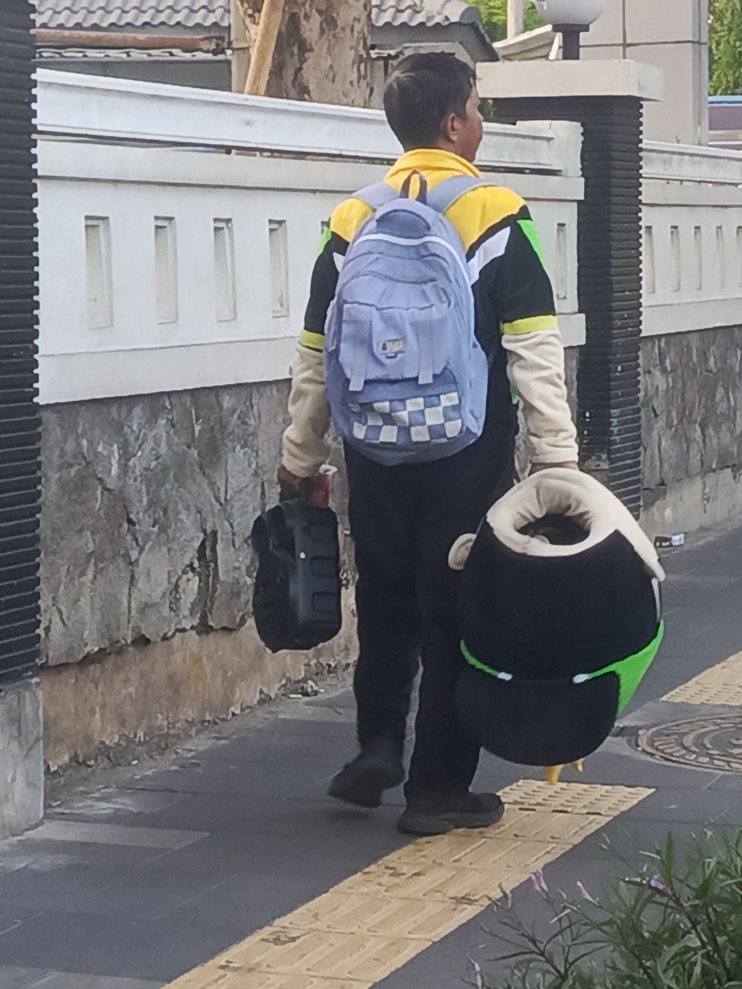 A person is walking on a sidewalk in Surabaya, Indonesia, carrying a light blue backpack, a black carry-on suitcase, and a large, round, stuffed toy with black, green, and white accents. The person is wearing a black shirt with yellow and green accents and black pants. The background shows a stone wall and a white fence. The sidewalk has tactile paving.