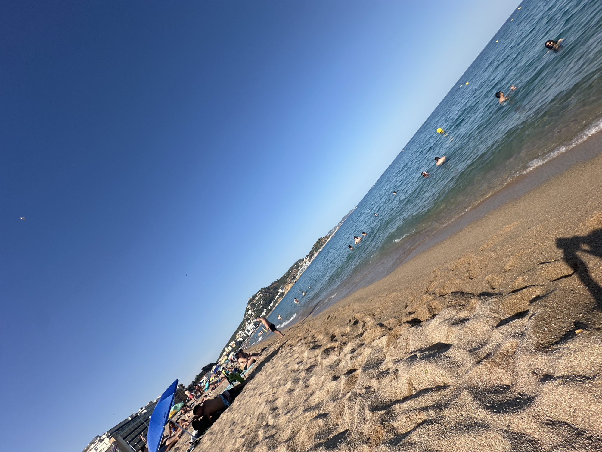 The image depicts a crowded beach scene in Blanes, Spain, during daylight hours. The perspective is from a low angle, looking diagonally across the sandy beach towards the sea. The sand is light brown with visible footprints and small pebbles. On the left side of the frame, a blue beach umbrella is prominent, with several people seated or lying on towels beneath it and around it. Some individuals are partially visible, wearing swimwear. In the background, a strip of buildings lines the coast, indicating an urban area adjacent to the beach. The sea is a vibrant blue, with numerous people swimming and wading in the water. Yellow buoys are scattered in the sea, marking a swimming area. The sky is clear and bright blue. The overall impression is a lively summer day at a popular coastal resort.