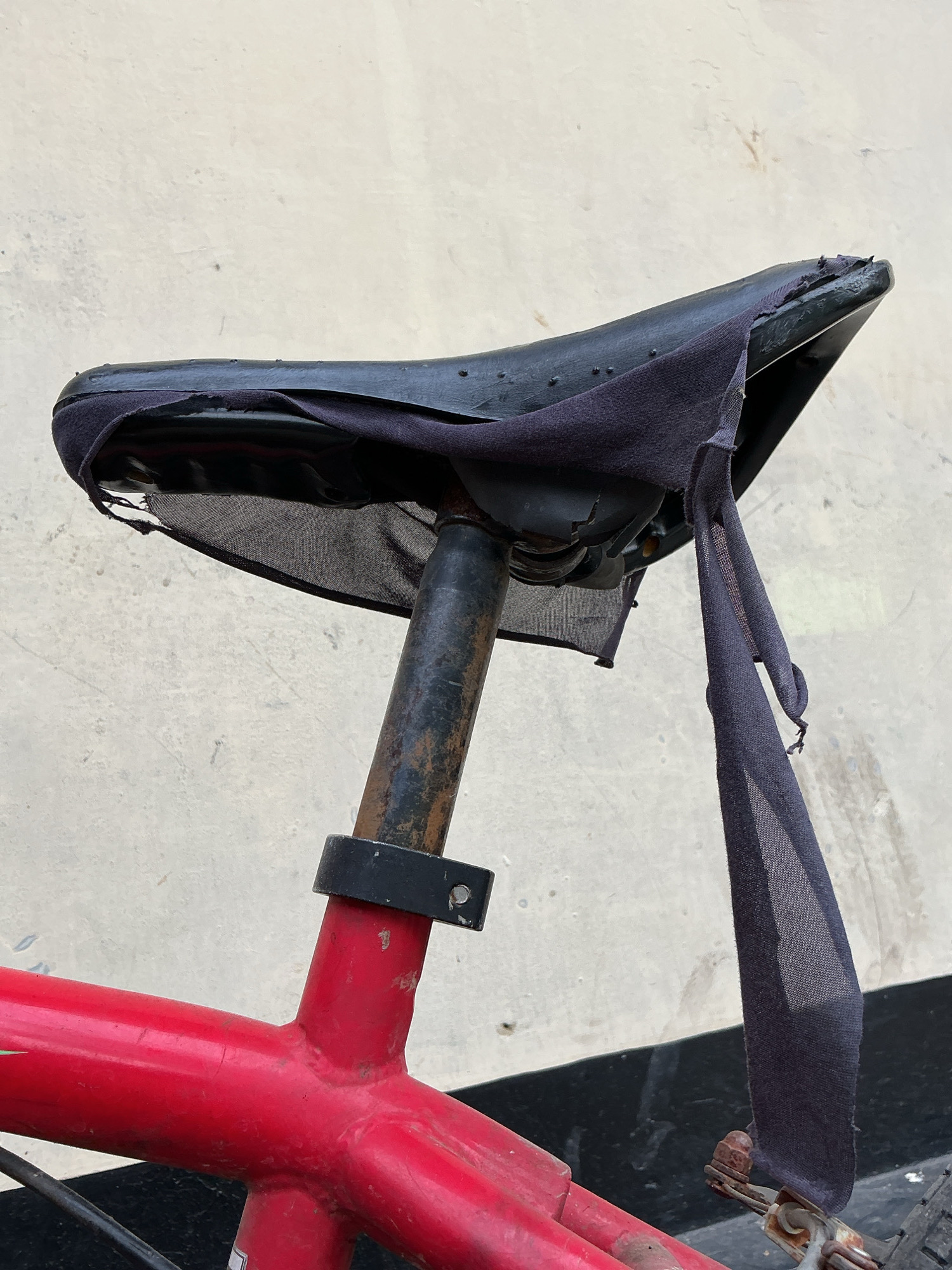 The image shows a close-up of a worn and torn bicycle seat. The seat is black and appears to be made of synthetic material, with some holes visible in the surface. A dark-colored fabric is draped over the seat, with one side hanging down and torn into strips. The seat post is rusted and metal, attached to the red frame of the bicycle. The background is a textured, light-colored wall, possibly concrete. The scene is set outdoors, with a dark surface visible at the bottom of the frame. No people are present in the image. The image quality is clear, with good lighting. The scene is likely in a public area or an outdoor storage space. The specific location in Magelang, Indonesia, cannot be determined from the visual information provided.