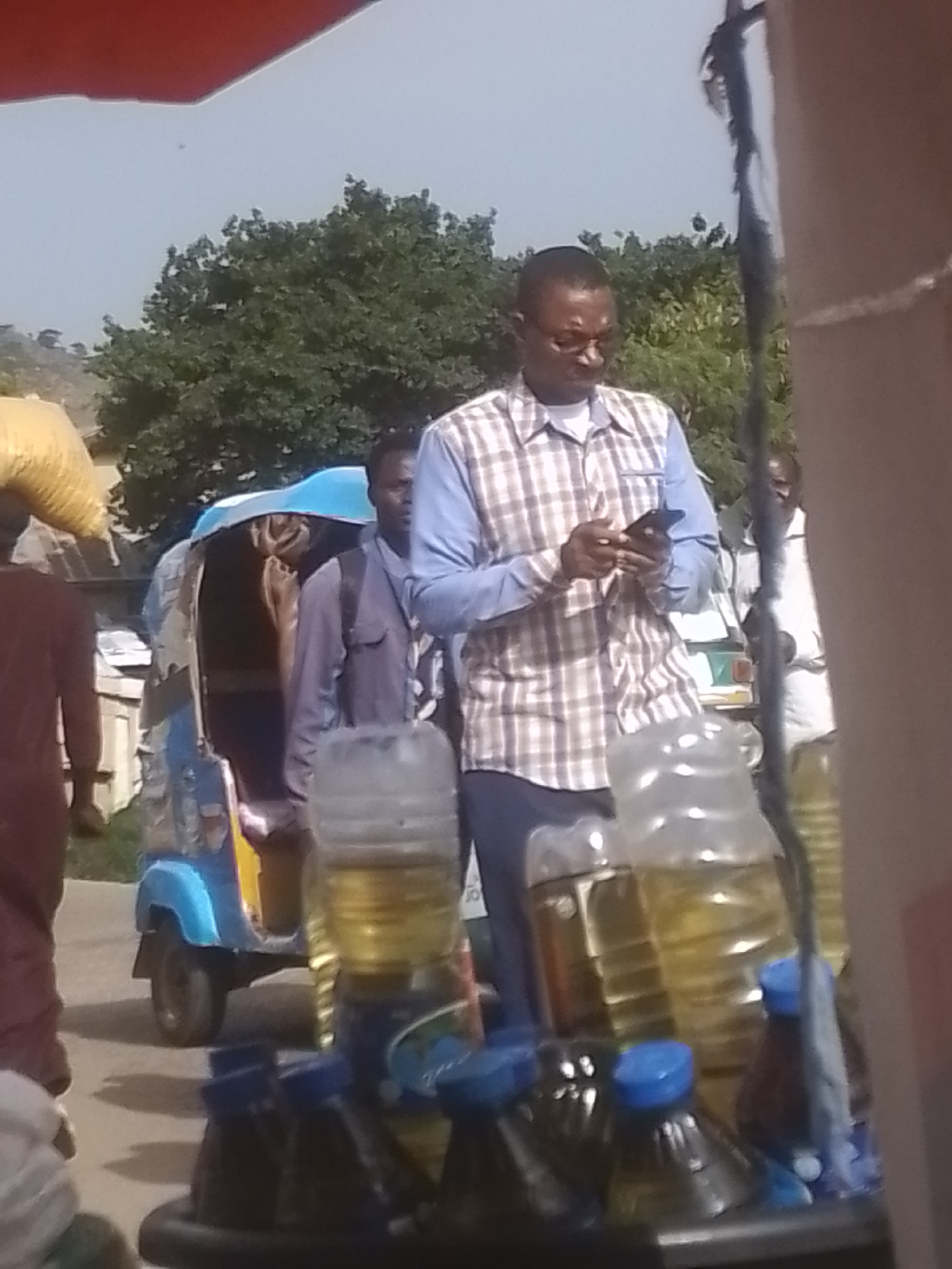 The image depicts an outdoor scene with several people and objects. In the foreground, there are several bottles filled with a yellow liquid, possibly oil, with blue caps. One man, wearing glasses and a plaid shirt over a white undershirt, is looking down at his phone. Behind him, a blue and yellow auto-rickshaw is visible, with a person inside. Further in the background, other individuals are present, and a person in the far left is carrying a large yellow sack on their head. Trees and a building are visible in the distance. The lighting suggests it is daytime.