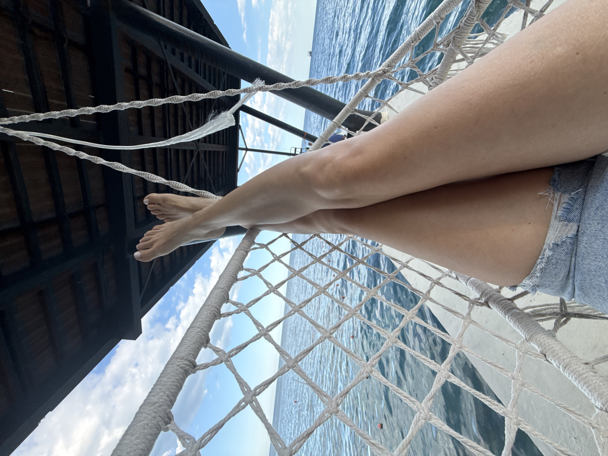 The image shows a person's legs and feet, crossed at the ankles, resting on a net hammock. The hammock is suspended from a wooden structure above, which appears to be part of a pier or boardwalk. The background consists of the calm, blue sea and a cloudy sky. In the distance, a ship is visible on the horizon. The overall scene suggests a relaxed, outdoor setting, possibly a vacation spot. The location is identified as Yalta, Ukraine.