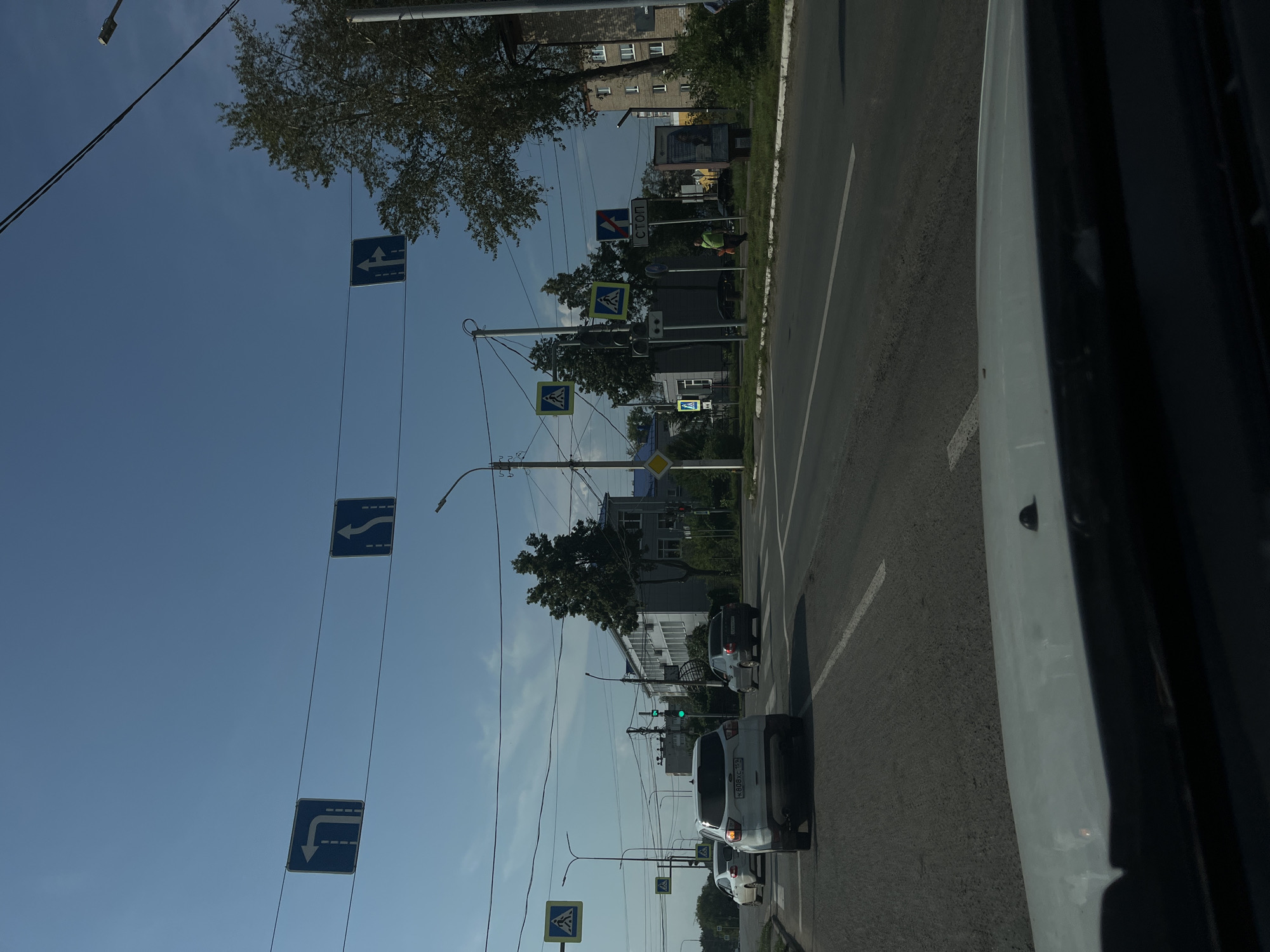 This is a forward-facing, eye-level, medium shot of a street scene in Chaikovsky, Russia, taken from inside a vehicle. The sky is clear and blue, with a few wispy clouds. Power lines and traffic lights are visible, strung across the road.
Several traffic signs are present:
- Two blue rectangular signs with white arrows indicating directions are suspended over the road. One shows a left turn only, and the other indicates a straight or right turn.
- A blue square sign with a white pedestrian crossing symbol is on the left side of the road.
- Another similar pedestrian crossing sign is visible further down the road on the left.
- A blue rectangular sign with a white arrow indicating a straight or left turn is visible to the right of the road.
- A blue rectangular sign with a white arrow indicating a straight direction is further down the road on the right.
- A blue and red rectangular sign with a white "STOP" text and a red cross shape is visible to the right.
Several cars are on the road. A white SUV is in the foreground, facing away from the camera. To its left, a white sedan is also facing away. Further down the road, a dark sedan is visible.
Buildings line the right side of the street. The closest one is a multi-story building with a light-colored facade and several windows. Further down, another building is visible. Trees with green foliage are also present.
A person wearing a green vest and holding a red object is walking on the sidewalk on the right side of the road. A pedestrian crossing is marked on the road. The overall scene depicts an ordinary urban intersection with typical traffic and street furniture.