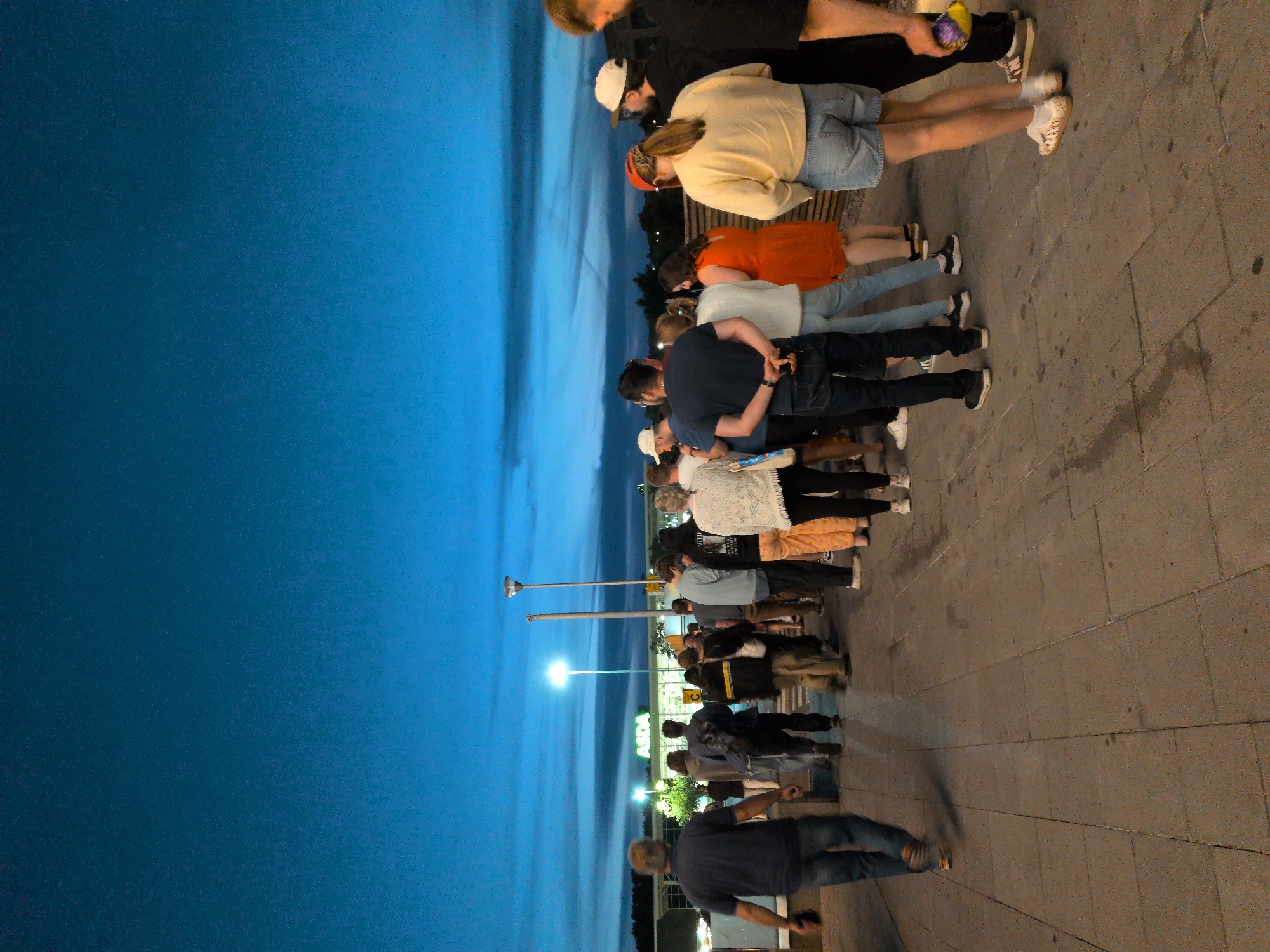 A group of people, appearing to be of various ages and genders, stand on a paved walkway outside a supermarket. The supermarket, identified by the "ASDA" sign, is visible in the background. The sky is a deep twilight blue, suggesting it is evening. The people are facing away from the camera, towards the supermarket entrance. Some individuals are wearing casual clothing such as t-shirts, jeans, and shorts. One person is holding a bag. The lighting suggests artificial lights from the supermarket and streetlights are illuminating the scene. The overall impression is that the group is waiting to enter the supermarket or perhaps leaving after it closed. The location is Asda, Telford, United Kingdom.