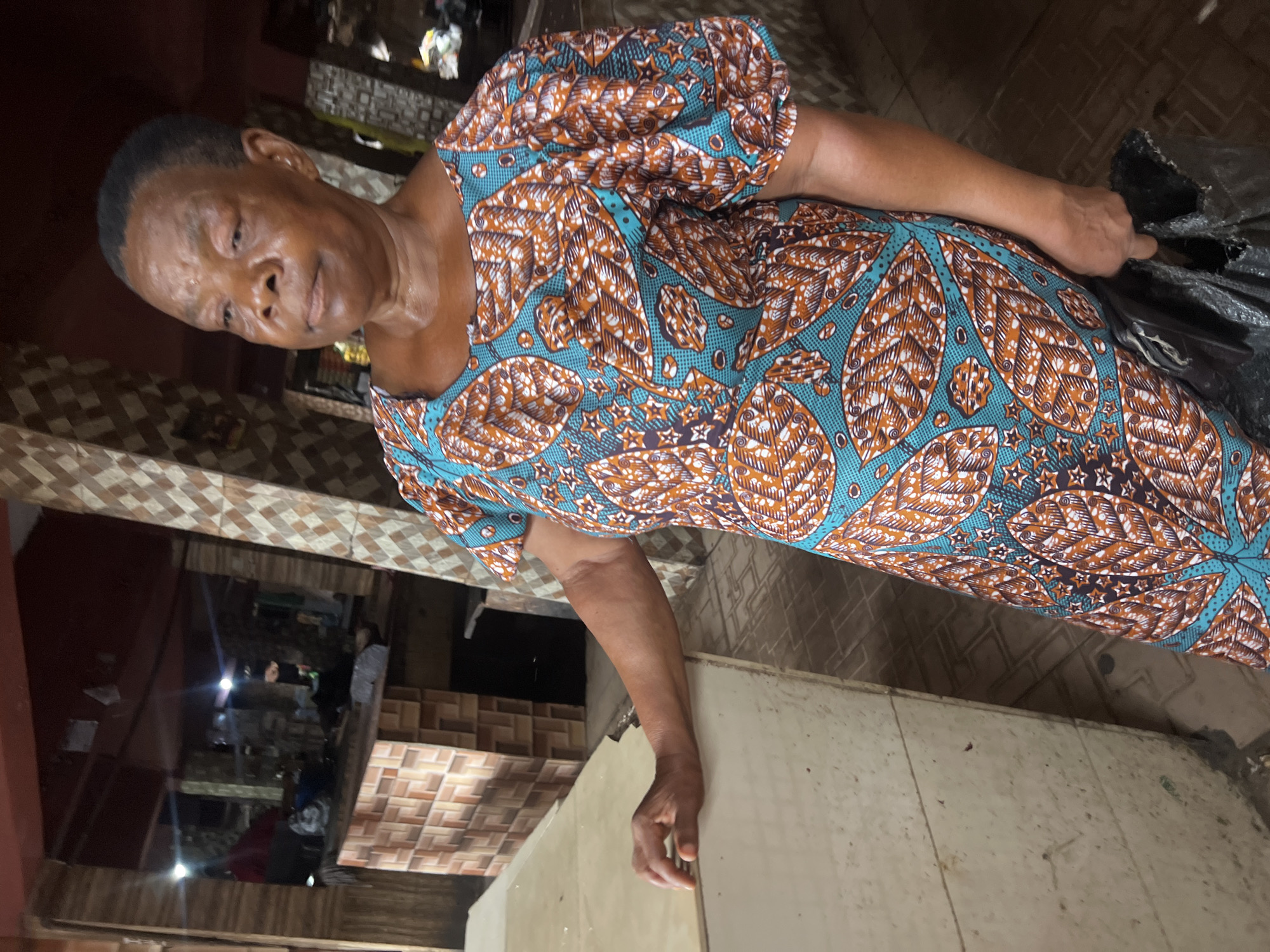 An elderly African woman stands within a building in Nkpor, Nigeria. She wears a vibrant dress with teal, orange, and brown leaf patterns. Her short, dark hair is neatly styled. The building's interior is visible, showcasing tiled walls and a glimpse of other people in the background. One of her hands rests on a light-colored surface, while the other seems to be holding onto something black.