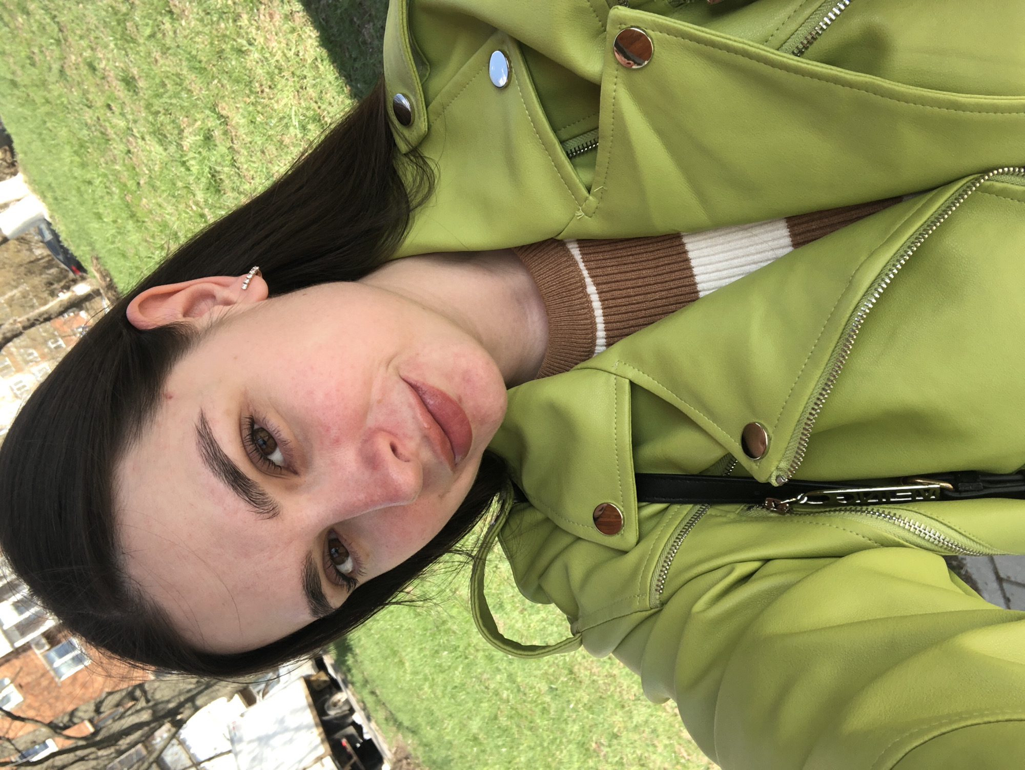 A woman is taking a selfie outdoors on a sunny day. She has dark hair, fair skin, and is wearing a light green leather jacket with metal buttons and zippers. Underneath, she wears a brown and white striped top. She has an earring in her left ear. The background shows a grassy area with buildings visible in the distance. Given the context of Votkinsk, Russia, the buildings could be residential or commercial structures typical of the city. The selfie style suggests a casual, personal moment captured by the woman.