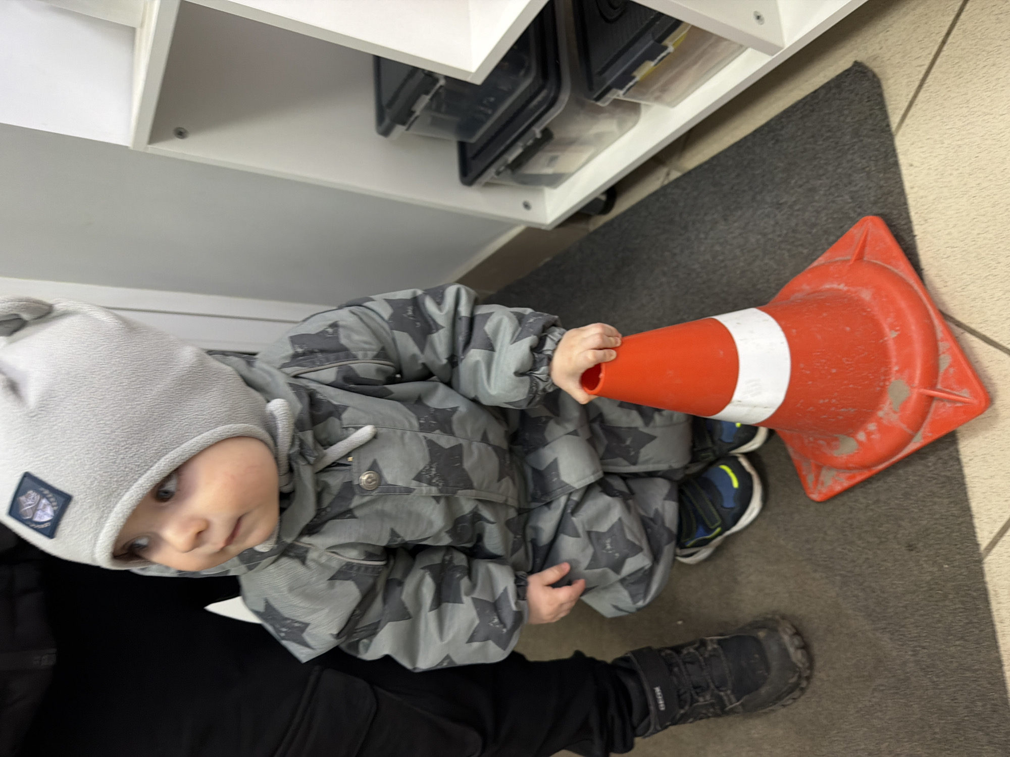 A young child, likely a toddler, is captured from an overhead perspective, sitting or kneeling on a dark grey floor mat. The child wears a light grey fleece hat with a dark square patch featuring a white logo, possibly "WILD," and a padded grey and dark grey star-patterned winter suit. Their blue and yellow sneakers are visible on the mat. The child holds the narrow end of a red and white traffic cone, which rests on its side on the mat. The child's gaze is directed upwards and slightly to the right, showing a curious expression.
Next to the child, the lower leg and black shoe of an adult are visible, suggesting the adult is accompanying or supervising the child. The adult's black shoe has the brand name "GEOX" faintly visible on it. In the background, a white shelving unit with black and clear storage containers is visible. The floor transitions from the grey mat to light-colored tiled flooring on the right side of the image. The setting appears to be indoors, possibly a public or commercial space in Votkinsk, Russia. The child's warm attire suggests cold weather outside. The time of day is not discernible from the lighting.