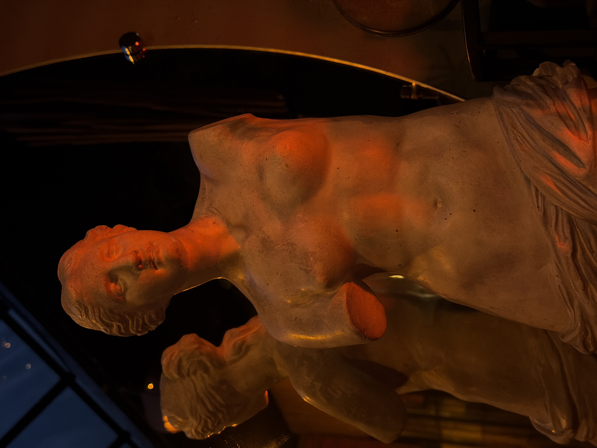 A close-up, slightly canted, low-angle shot depicts a partially fragmented female statue with a reflection on a dark surface, likely glass or polished stone. The statue is illuminated with warm, possibly artificial light, casting an orange hue on the white material of the sculpture. The statue is headless, armless, and appears to be a torso draped with fabric on one side. The reflection shows a distorted view of the face and shoulders.
In the background, there are hints of a dark interior space with a window showing a faint blue or dark sky. There is a small, indistinct, colorful light fixture suspended from the ceiling. A small part of the restaurant's interior design is visible. The location of this image is Yerevan, Armenia, specifically at the restaurant located at 12 Pushkin Street.