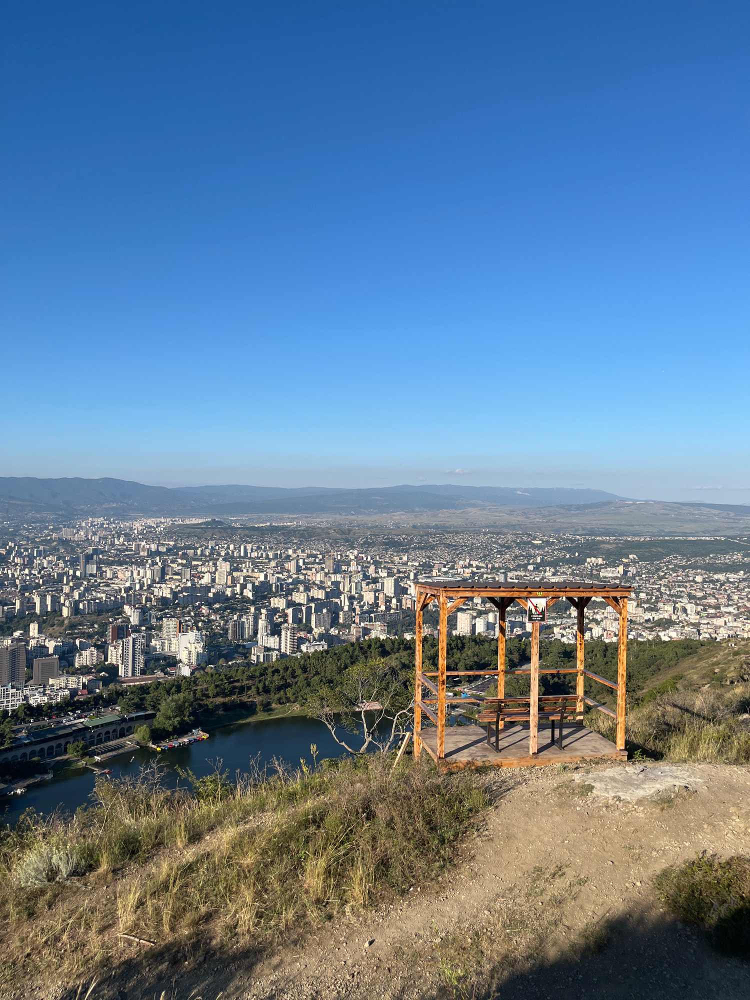 The image captures a panoramic view of Tbilisi, Georgia, from an elevated vantage point. In the foreground, a wooden gazebo with a picnic table and benches sits on a grassy hillside. Beyond the gazebo, a body of water, likely a lake or reservoir, is visible, bordered by lush green trees. The city sprawls across the landscape in the middle ground, characterized by a dense collection of buildings of varying heights, predominantly white and grey, set against a backdrop of distant, hazy mountains under a clear blue sky. The scene appears to be set during the daytime, with ample sunlight casting shadows on the terrain. There are no people visible in the frame.