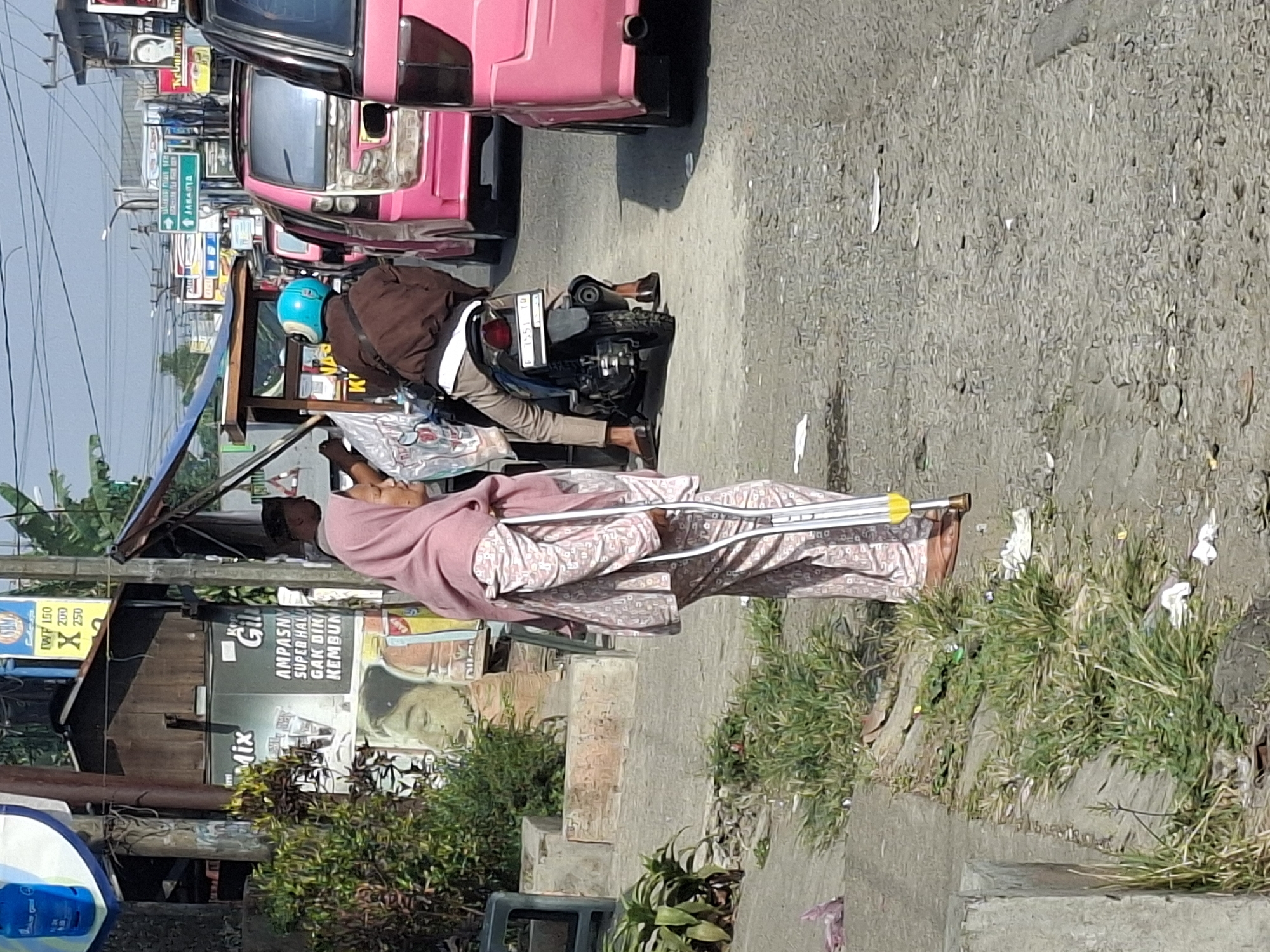 In the city of Sukaraja, Indonesia, a woman wearing a pink headscarf and a patterned pink outfit stands on a sidewalk, leaning on a white crutch with a yellow handle. She is facing towards the left of the frame. Behind her, a man in a brown jacket and a blue helmet rides a motorcycle. In the background, several pink vehicles are lined up, suggesting a busy street. There are also signs and stalls that indicate a market or roadside shops. The ground is paved with asphalt and gravel, with some patches of grass and debris visible along the edge of the sidewalk. The sky is clear and bright, indicating it is daytime.