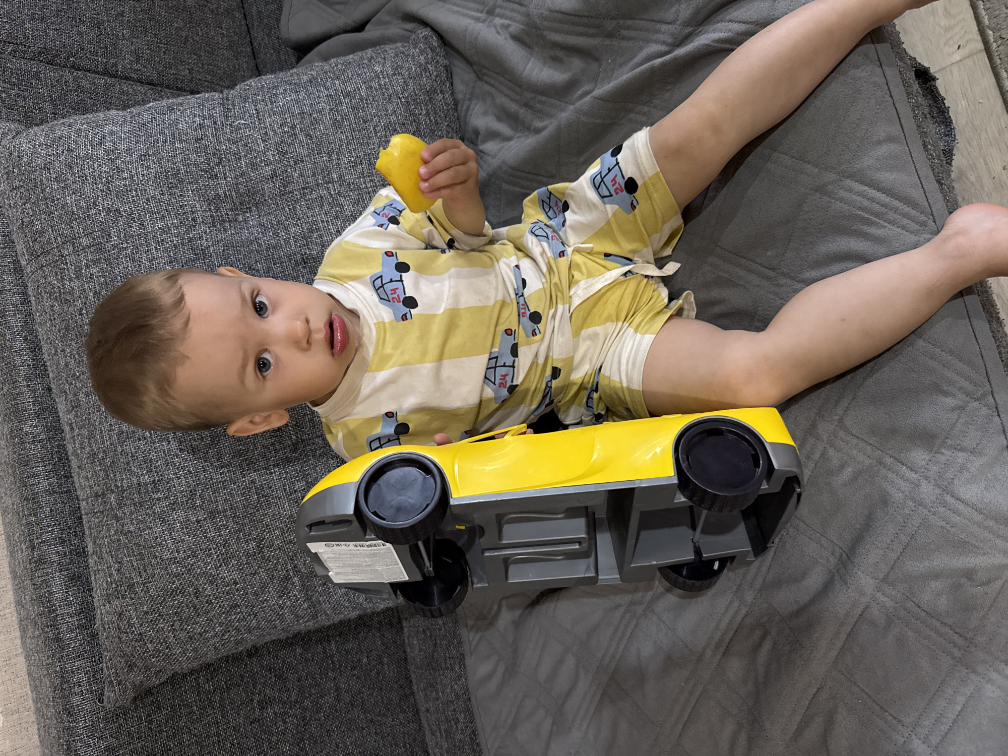 This image features a young child, likely a toddler, lying on a grey fabric sofa or bed in an indoor setting. The child has light brown hair and direct, wide blue eyes, looking towards the camera with an open mouth. They are wearing a long-sleeved top and shorts in yellow and white stripes, patterned with blue cartoon trucks, some of which display the number "24". In their right hand, the child holds a piece of yellow citrus fruit, possibly a lemon.
Near the child's feet, a large yellow and grey toy car is positioned upside down, revealing its underside and black wheels. The grey fabric surface beneath the child and the car appears to be a quilted blanket or throw. A glimpse of a lighter-colored rug or carpet and a wooden floor is visible along the right edge of the frame. The overall scene suggests a relaxed and playful moment in a home environment.
Notable details include the child's curious and direct gaze, and the details on their clothing. On the underside of the toy car, a white sticker with a barcode and some small, indistinct text is visible. The scene is set indoors, likely a living room or bedroom, in Шебунино, Russia.