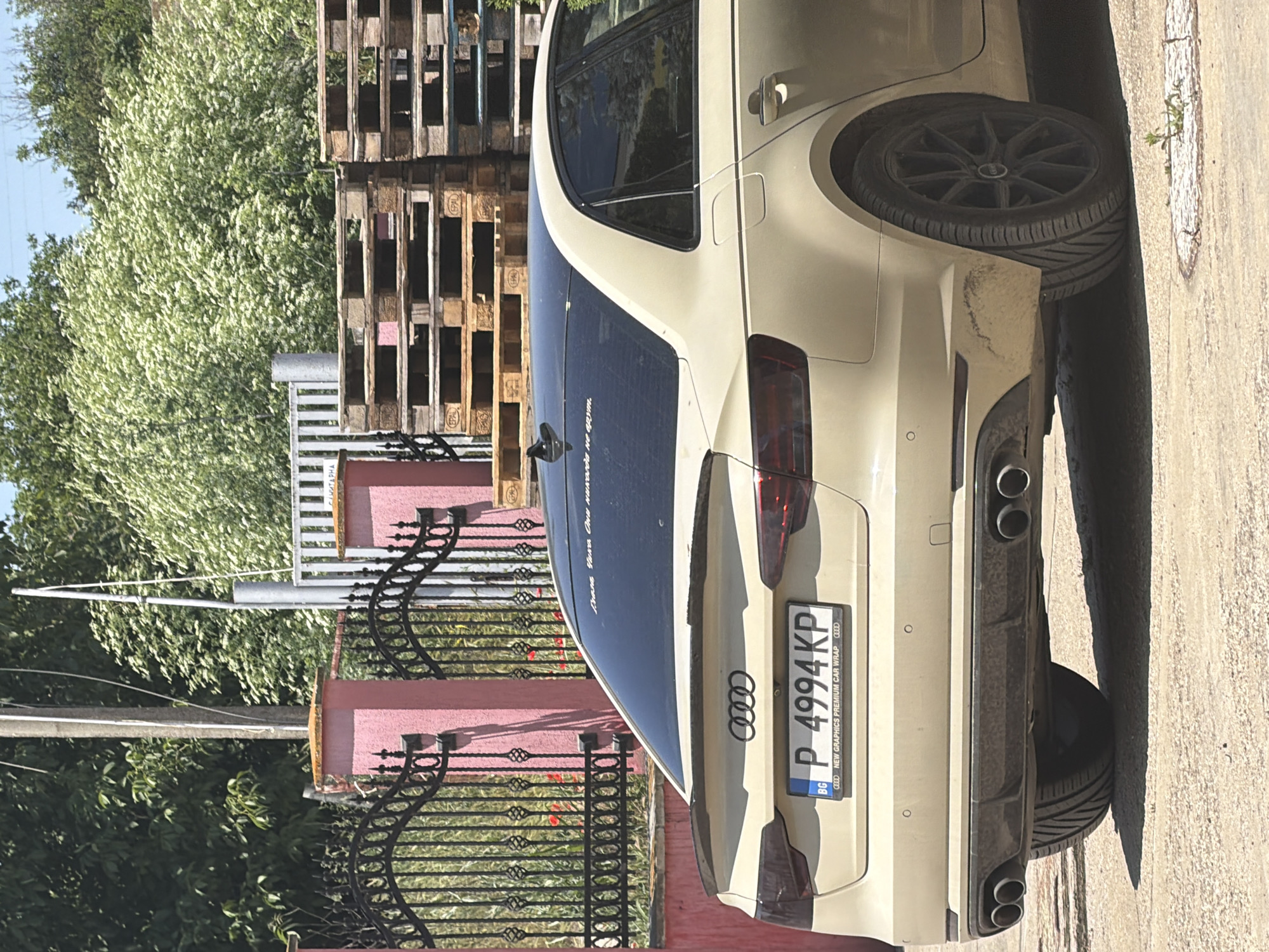 A beige Audi sedan is parked on a gravel surface. The car has a black roof and tinted rear windows. A spoiler is attached to the trunk. The license plate is visible and reads "P 4994KP BG". Text below the license plate says "NEW GRAPHICS PREMIUM CAR WRAP". There is a sticker on the rear window. The car is positioned in front of a pink wall with a black metal fence. Behind the fence are trees and a stack of wooden pallets. The scene appears to be in Dolno Ablanovo, Bulgaria, as indicated by the license plate information. The weather is sunny.