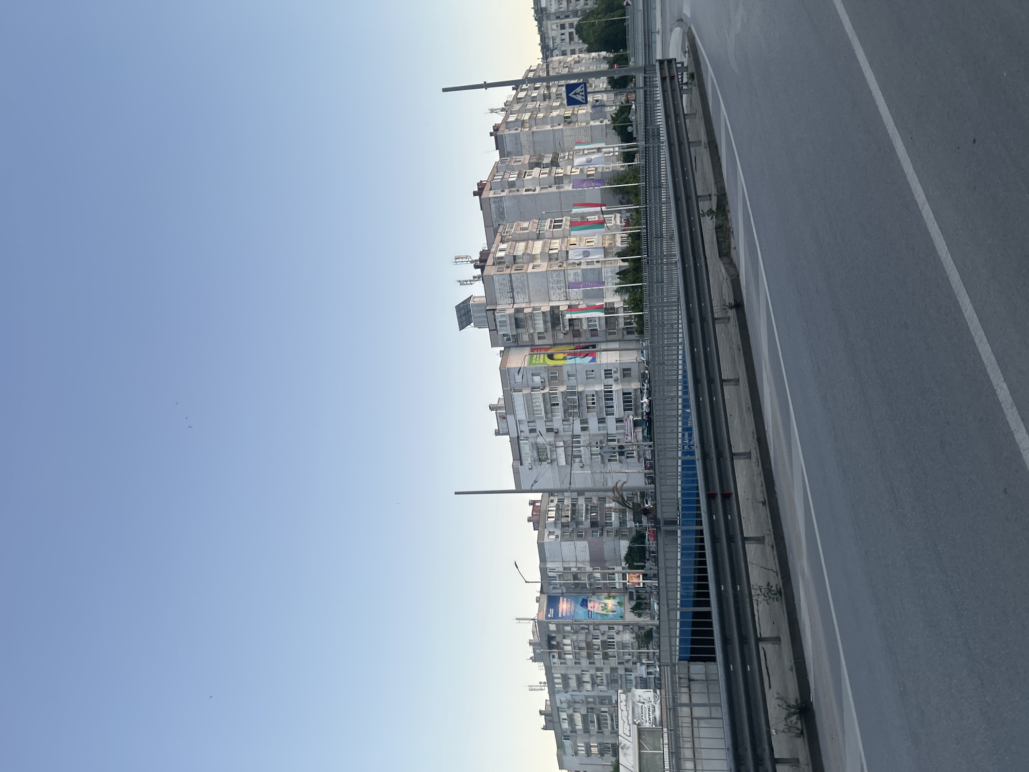 The image shows a street view in Varna, Bulgaria, featuring a row of multi-story residential buildings under a clear blue sky. A highway overpass with a metal guardrail runs in the foreground, with a paved road visible below and to the right. The buildings are constructed in a typical Soviet-era architectural style, with light-colored facades, numerous windows, and balconies. Several flags are displayed in front of the buildings, including the Bulgarian flag, as well as flags of other nations or organizations. Some of the buildings have visible antennas and solar panels on their roofs. There are no people clearly visible in the frame. The overall impression is that of a residential district in a city, likely during daytime or early evening given the light.