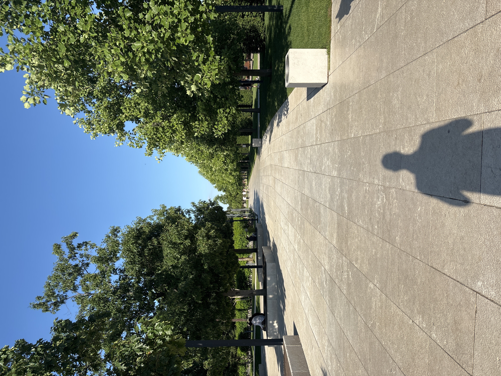 The image depicts a sunny day in a park, characterized by a wide, paved walkway lined with trees on both sides. The walkway is constructed of light-colored, rectangular tiles laid in a grid pattern. On the left side of the walkway, there are several individuals seated on benches or the ground beneath the trees. The trees are lush and green, casting shadows on the walkway. Several modern, cube-shaped planters are visible along the path. In the foreground, the shadow of a person is cast on the walkway. In the background, the path recedes into the distance, lined with more trees and indicating the presence of other people walking. The sky is a clear, bright blue. This scene is located in the Park of 30th Anniversary of the Victory in Krasnodar, Russia.