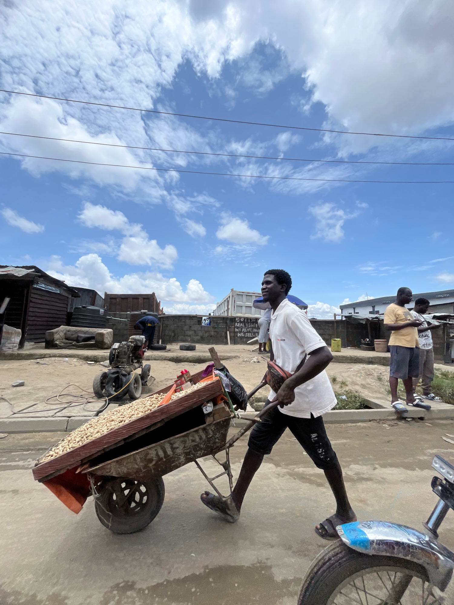 In Ado, Nigeria, a man in a white polo shirt and black shorts pushes a cart filled with peanuts along a paved street. The cart has wooden sides and metal framing, with a single wheel at the front. In the background, various structures are visible, including a dilapidated building with corrugated metal siding, a large industrial truck, and more modern buildings. Several other people are present, including two men standing near a wall, one holding a phone, and another in a yellow shirt. A small engine or generator is also parked on the ground. The sky is blue with scattered white clouds.