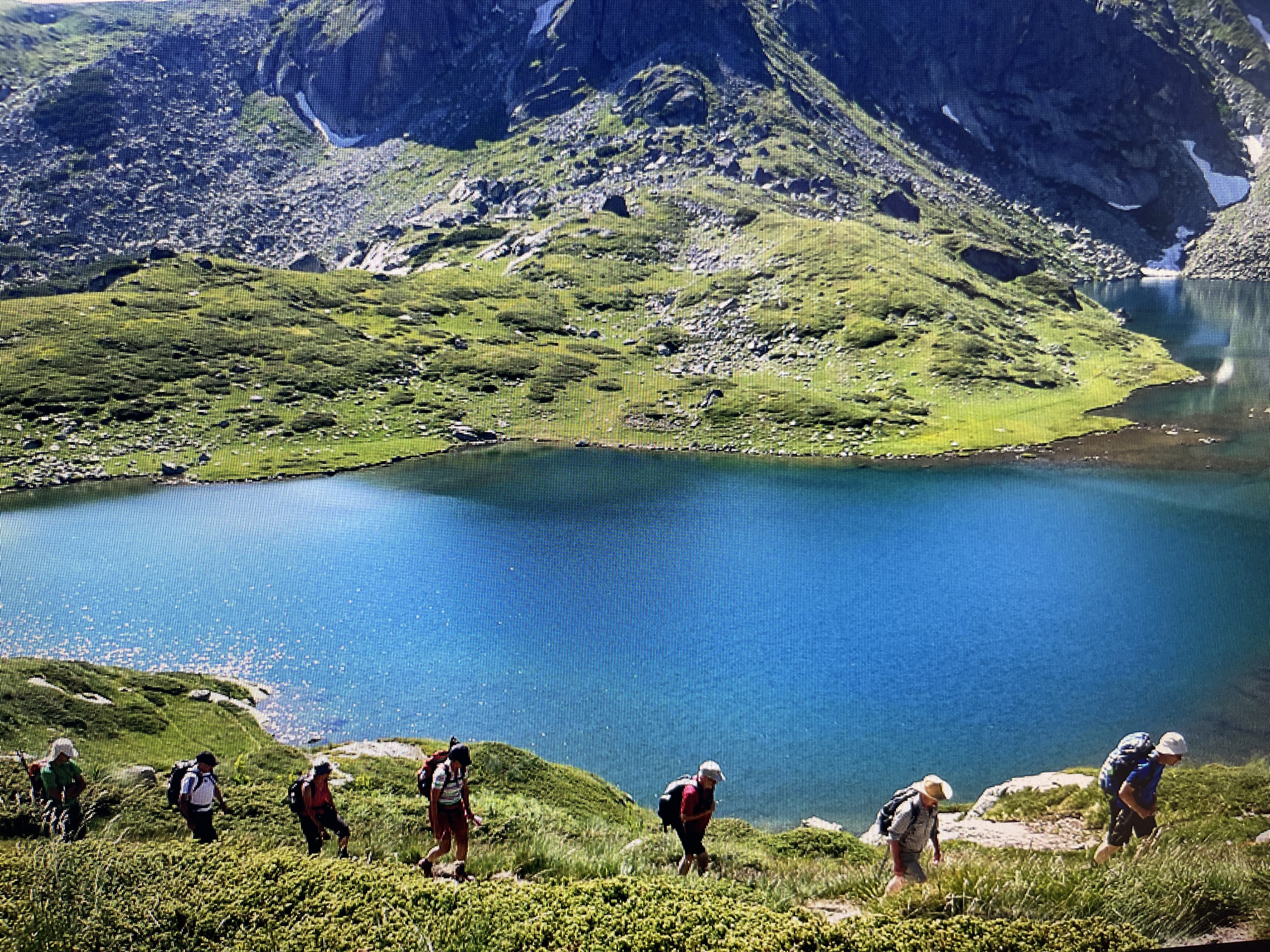 The image displays a group of seven hikers ascending a dirt path on a green, rocky hillside overlooking a vivid blue alpine lake under bright sunlight. In the foreground, dense green vegetation, primarily low shrubs and grasses, covers the terrain where the path is visible. The hikers, dressed in various colored hiking attire including shirts, shorts or pants, and hats, are all equipped with backpacks, and some appear to have hiking poles. They are spread out along the trail, moving from the lower left towards the upper right of the frame, indicating an uphill ascent.
The prominent feature in the midground is a large, brilliantly blue lake, its surface reflecting sunlight with numerous sparkling points. The immediate shoreline consists of green grassy areas transitioning to rocky or dirt patches. Beyond the lake, the background features imposing, dark grey mountainous terrain. These mountains exhibit steep, rocky slopes interspersed with patches of green vegetation and visible snowfields, particularly in shaded areas and higher reaches. Another smaller body of water is discernible nestled higher within the mountain landscape. The sky is clear and bright. The scene is located in the city of Varna, Bulgaria.