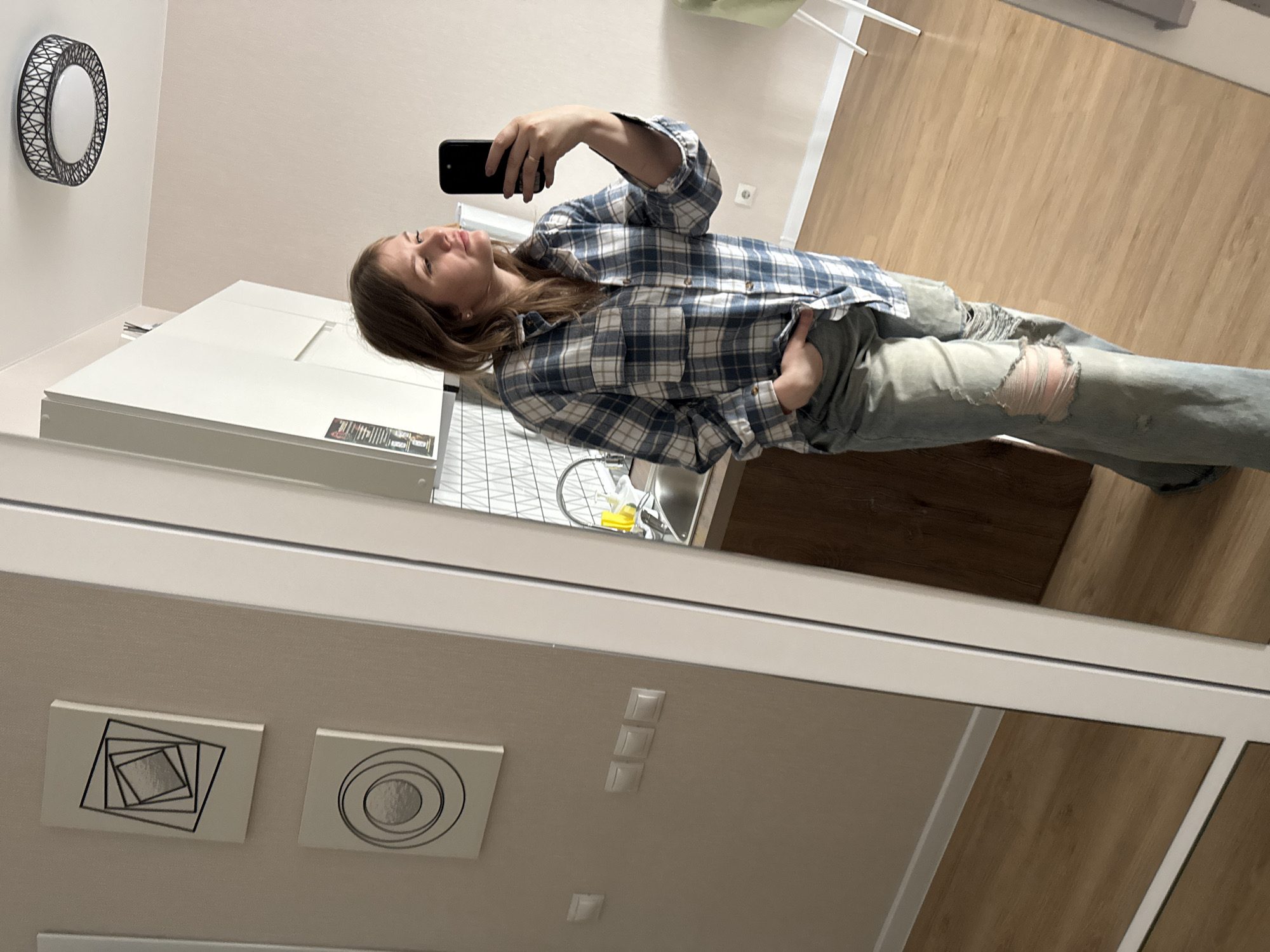 A woman is taking a selfie in a mirror. She is wearing a blue and white plaid shirt and ripped jeans. The background shows a kitchen area with white cabinets, a sink, and wooden floors. Two modern geometric wall art pieces are visible. The image was likely taken in Perm, Russia.