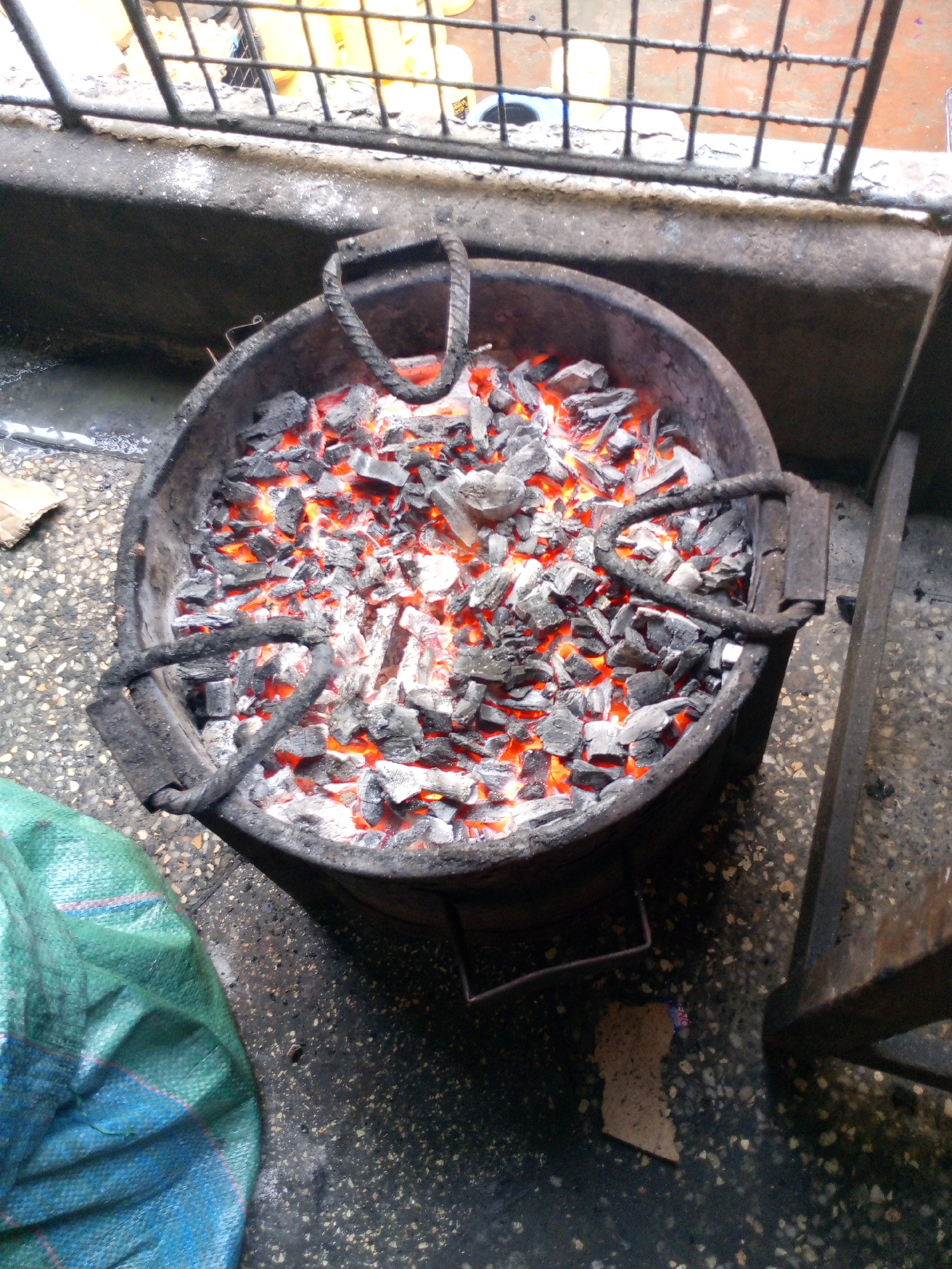 The image displays a dark, circular metal charcoal stove (jiko) filled with glowing red and orange charcoal embers and grey ash, viewed from an elevated perspective. The stove features two U-shaped rebar handles integrated into its rim and rests on a dark, granular concrete or asphalt surface. To the lower left, a section of a teal/green woven plastic sack is visible. To the lower right, an irregular piece of light brown cardboard lies on the ground, adjacent to a dark, vertical metal support. In the background, a low concrete wall separates the foreground from a dark metal grid fence. Behind the fence, multiple yellow plastic jerrycans are discernible. A dark, wet area is visible on the ground near the concrete wall. No individuals are present, and no explicit actions are depicted. The scene is located in an urban area of Nairobi, Kenya.