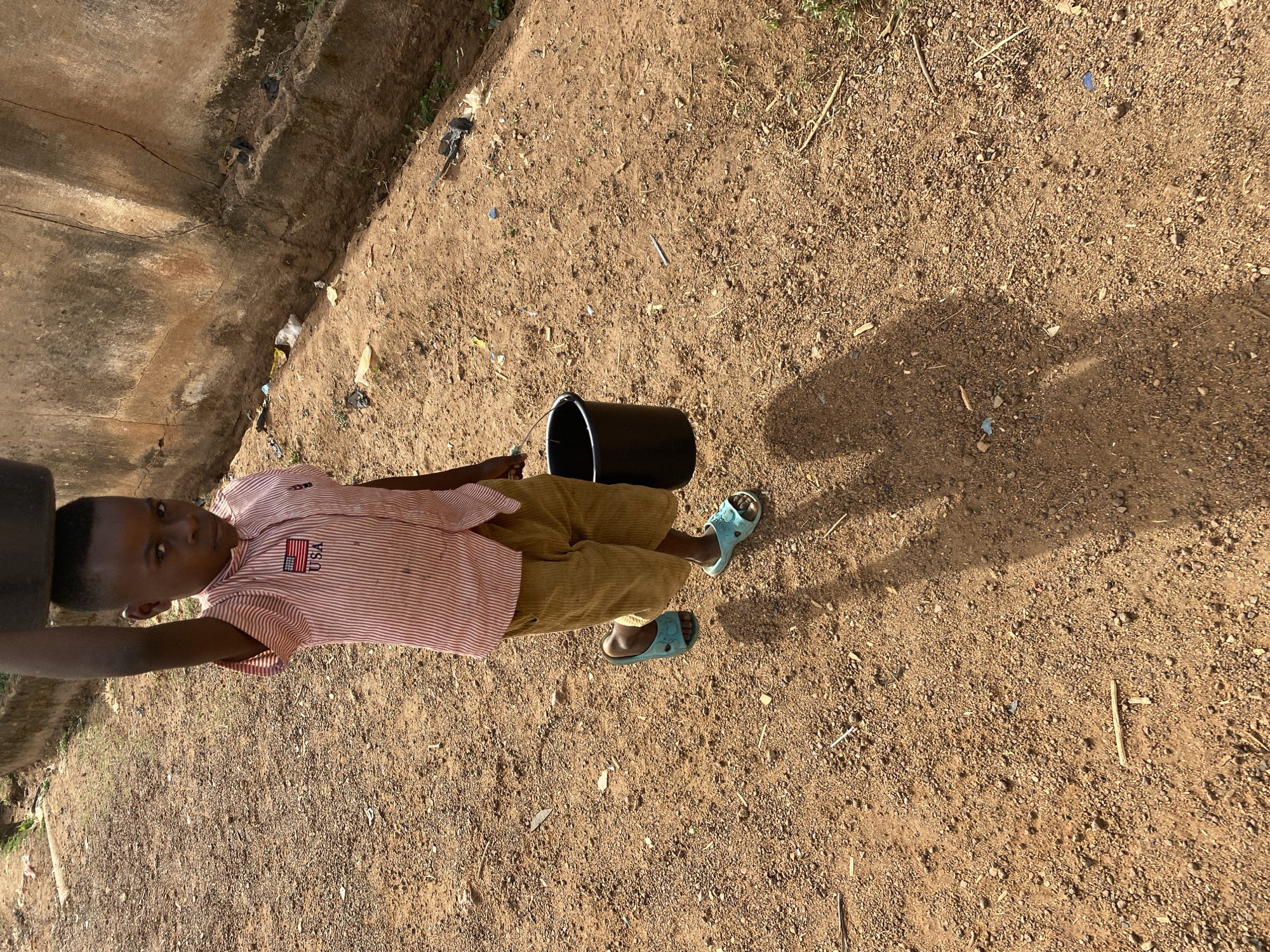 A young boy wearing a striped shirt with a USA flag patch, brown pants, and blue sandals stands on a dirt surface next to a black bucket. He is near a weathered, concrete structure. The location is Bukuru, Nigeria, as indicated by the metadata. The dirt surface is littered with small debris. The boy appears to be standing in sunlight, casting a shadow on the ground.