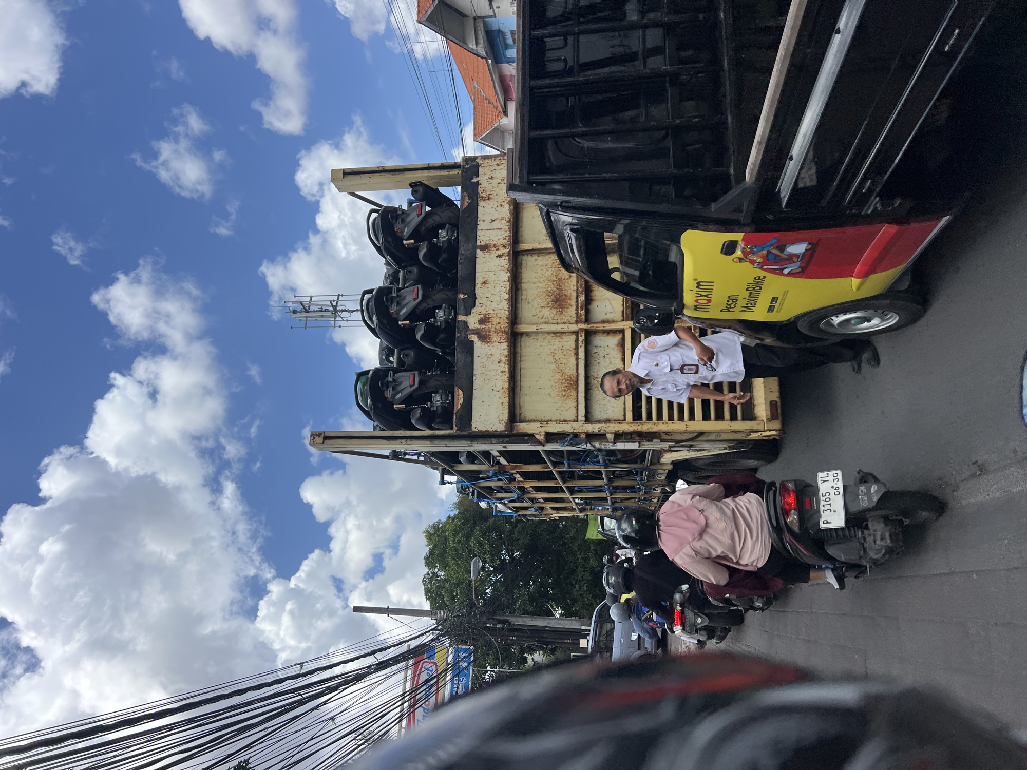In Denpasar, Indonesia, a traffic jam is captured on a cloudy day with a bright blue sky. The foreground shows the perspective of a person riding a motorcycle, with their helmet and vehicle's handlebars visible. Further ahead, several other motorcycles are in traffic, with riders wearing helmets and casual clothing. One rider, wearing a pink hoodie, is on a motorcycle with license plate "P 3165 YL 06.26". Adjacent to the traffic, a yellow truck is parked, displaying the Maxim logo and text that says "Pesan MaximBike", indicating a ride-sharing service. A man in a white uniform stands next to the truck. Behind the yellow truck, a larger, older truck is filled with several black motorcycles, stacked in its cargo bed. The background reveals buildings with red-tiled roofs and power lines overhead.