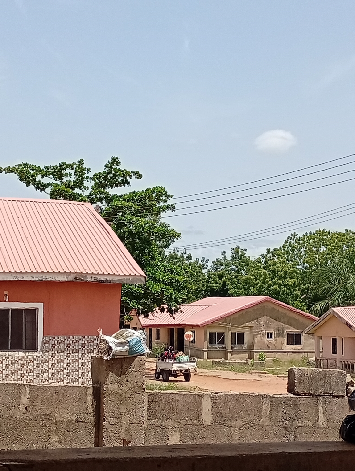 The image depicts a residential area under a slightly overcast sky. Several single-story houses with pink roofs are visible behind a concrete block wall. The houses appear to be of simple construction, with some showing signs of weathering or disrepair. A small motorized vehicle is positioned in the foreground with people inside. The area seems to be sparsely vegetated with various trees scattered throughout. Power lines stretch across the upper portion of the frame. The location is Unknown, Unknown.