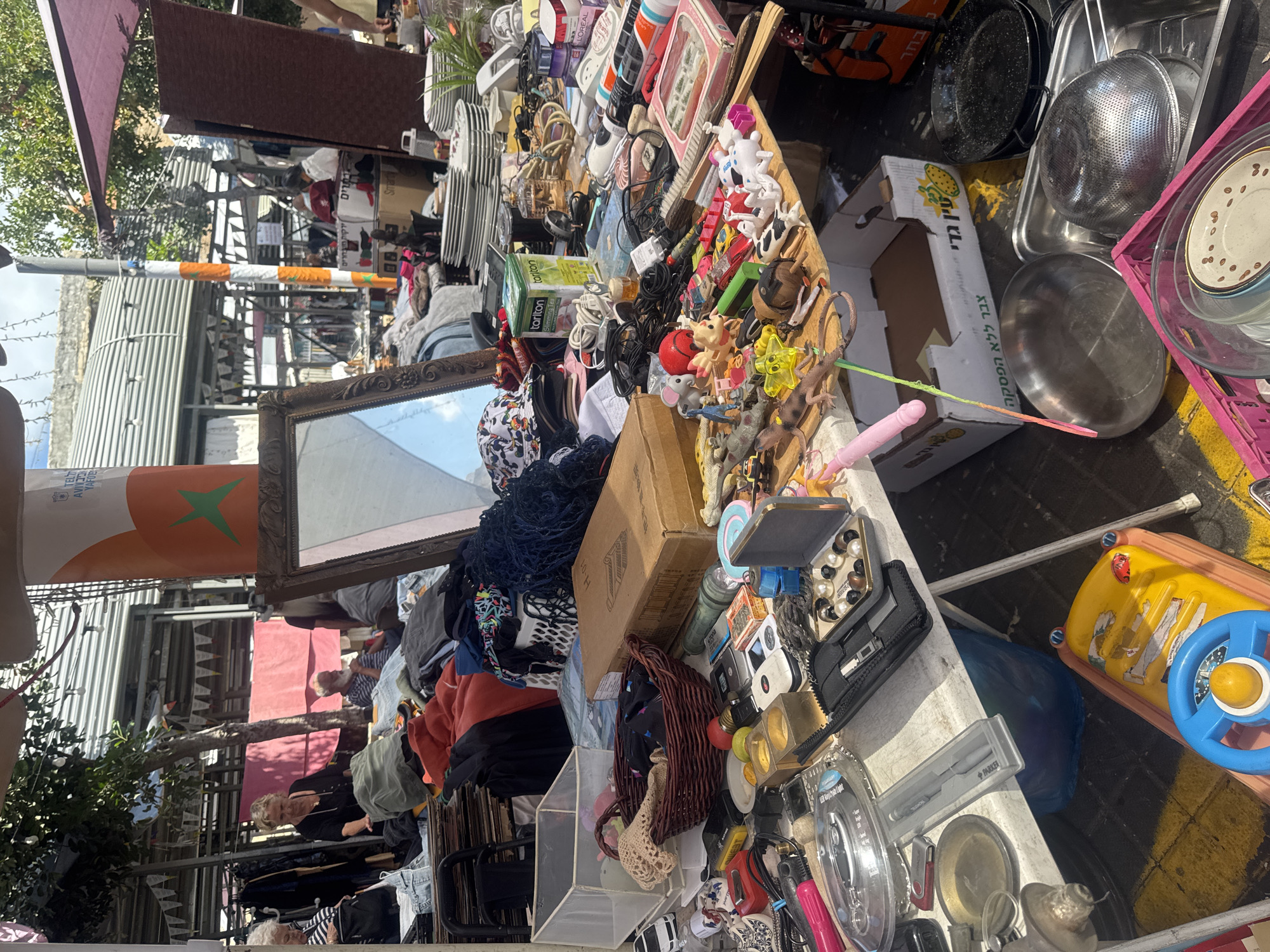 The image shows a crowded stall at a flea market in Tel-Aviv, Israel. The stall is covered with a variety of items, including clothing, housewares, toys, and electronics. There is a mirror propped up on the left side of the stall, and a sign in Hebrew. In the background, there are people browsing the market. A mix of metal containers, cookware, plates, and other tableware are scattered on the table. A children's riding toy is visible on the right.