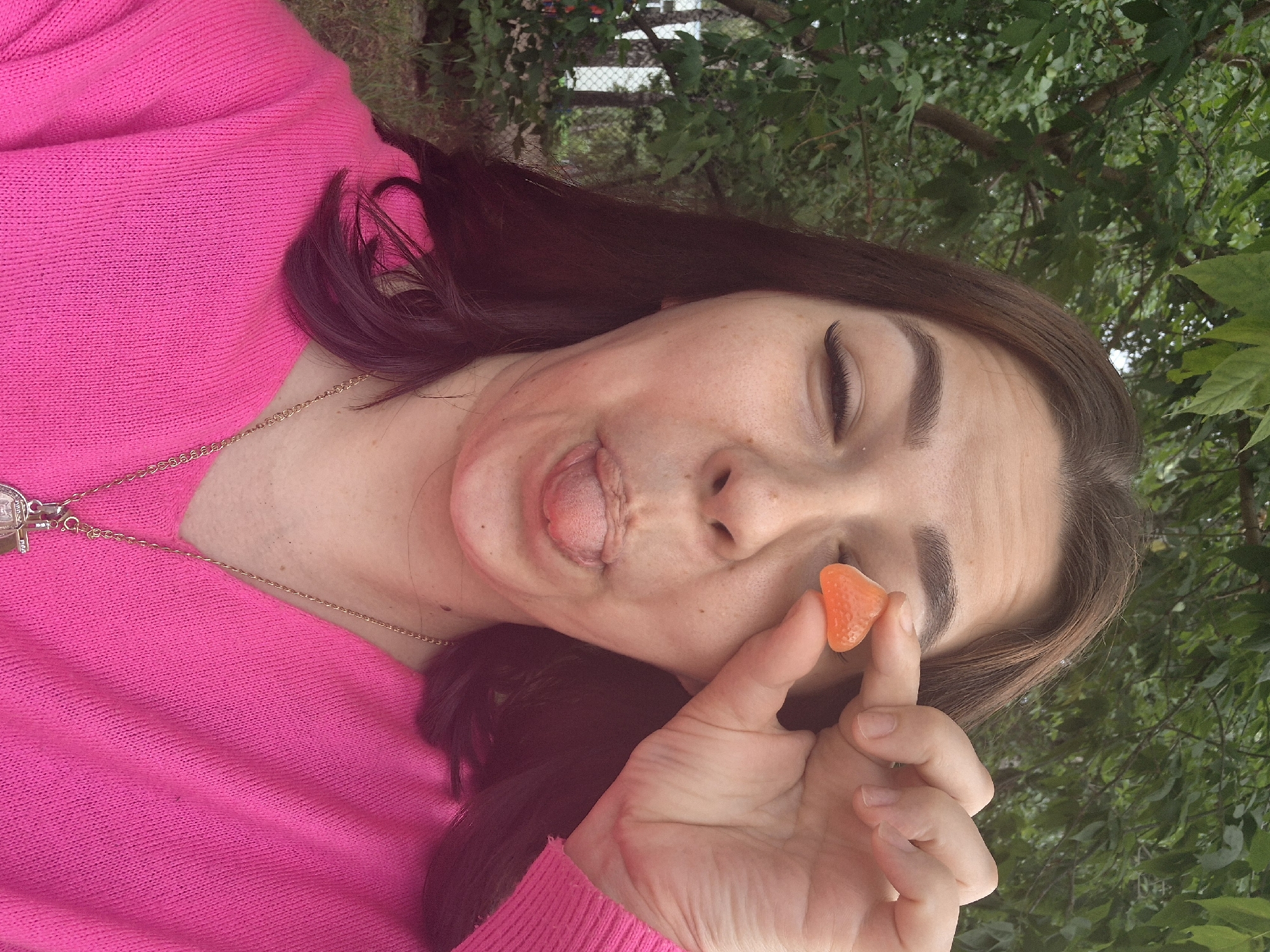 A woman with dark hair and light skin, wearing a bright pink sweater, is in an outdoor setting with green trees in the background. She is holding a triangular orange gummy candy in front of her right eye and sticking her tongue out. The scene appears to be in a park or backyard, and given the provided location context, it is likely in the city of Chaikovsky, Russia.