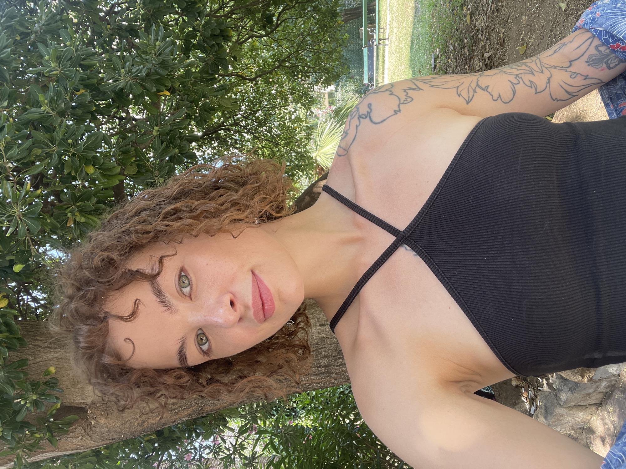 A young woman with curly brown hair and light skin is taking a selfie outdoors. She is wearing a black tank top and has a tattoo on her left arm. She is standing in front of a tree with green foliage and appears to be in a park or garden. The background shows more trees, green grass, and a park bench. The lighting suggests it is daytime and sunny. The location is identified as the city of Budva, Montenegro.