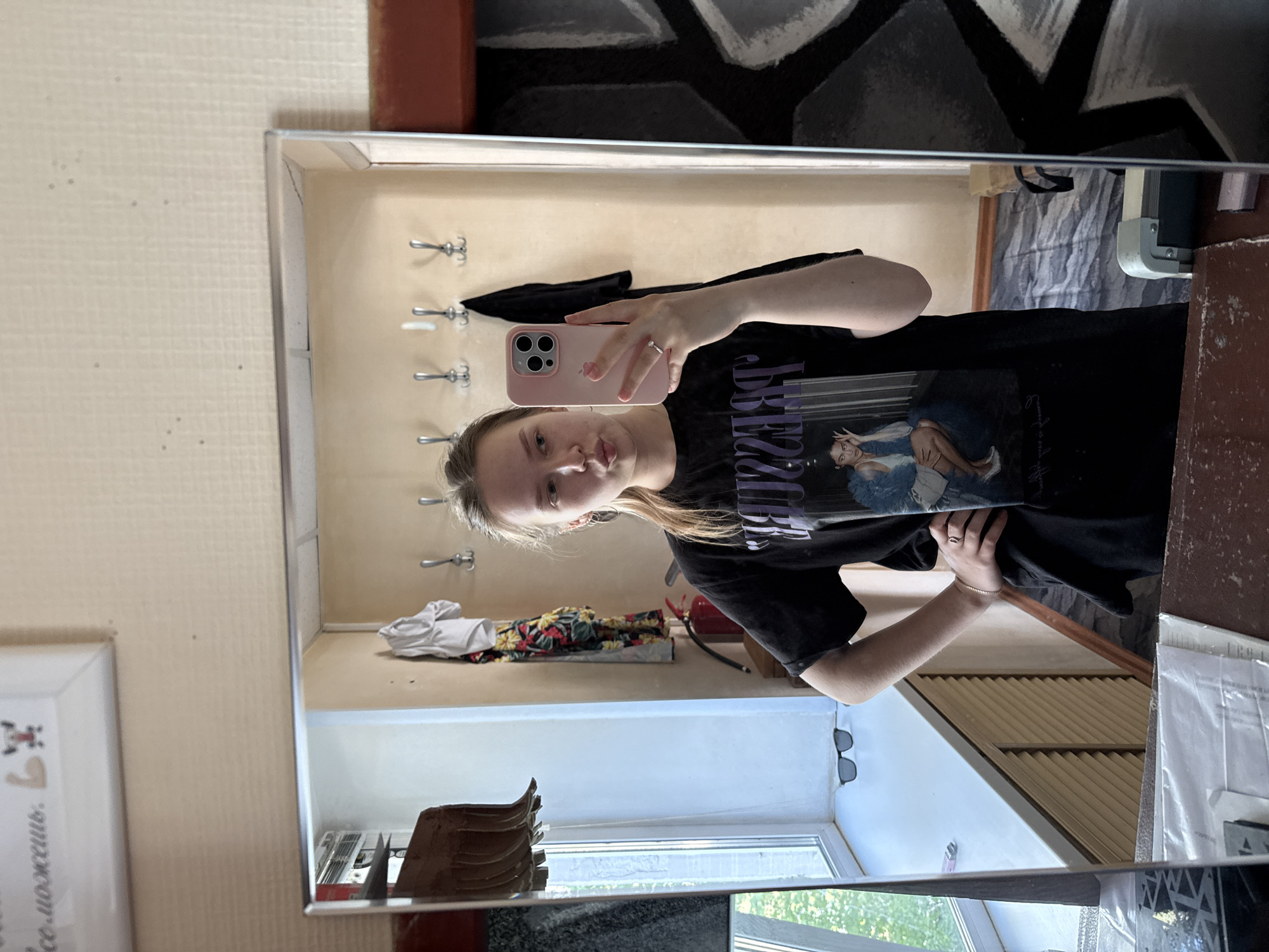 A young woman takes a selfie in a mirror. She is holding a pink smartphone and wearing a black t-shirt with a graphic print. She has light blonde hair. The mirror reflects a hallway with a coat rack, clothes, a window, and sunglasses on the windowsill. The location is likely Chaikovsky, Russia, based on context.